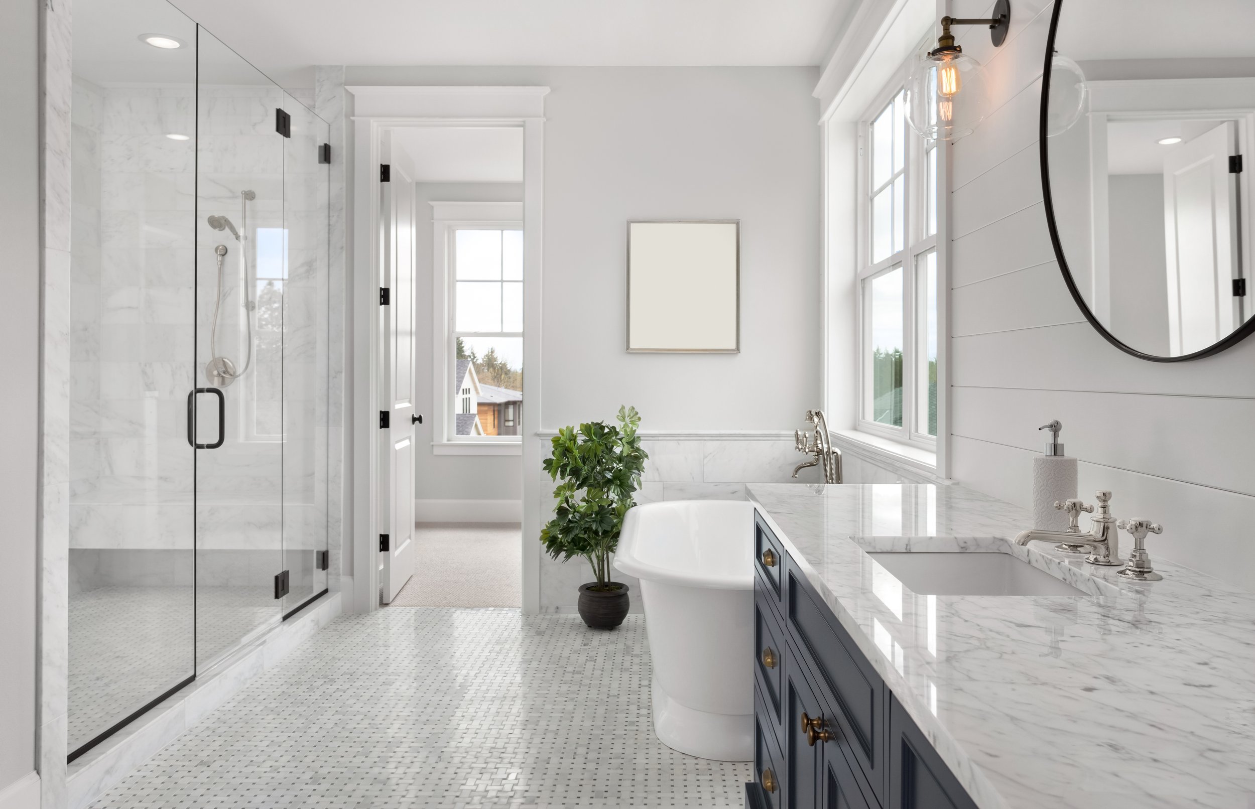 A bright, modern bathroom with a glass-enclosed shower, white wood-paneled walls, a white bathtub, large windows, and dark blue cabinets with gold hardware. There is a round mirror, a wall-mounted light, and a potted plant.