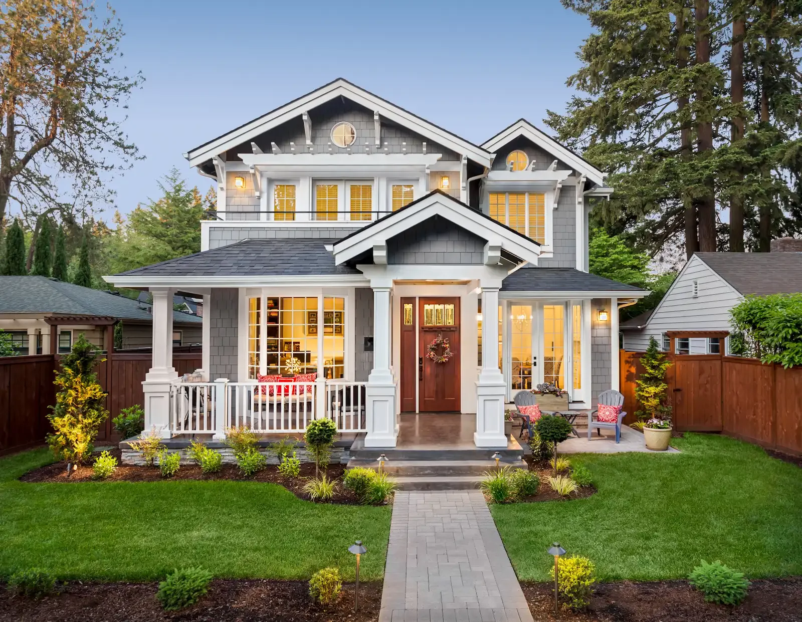 Front yard of a modern two-story house with a porch, lit windows, green lawn, small trees, and a walking path leading to the front steps.