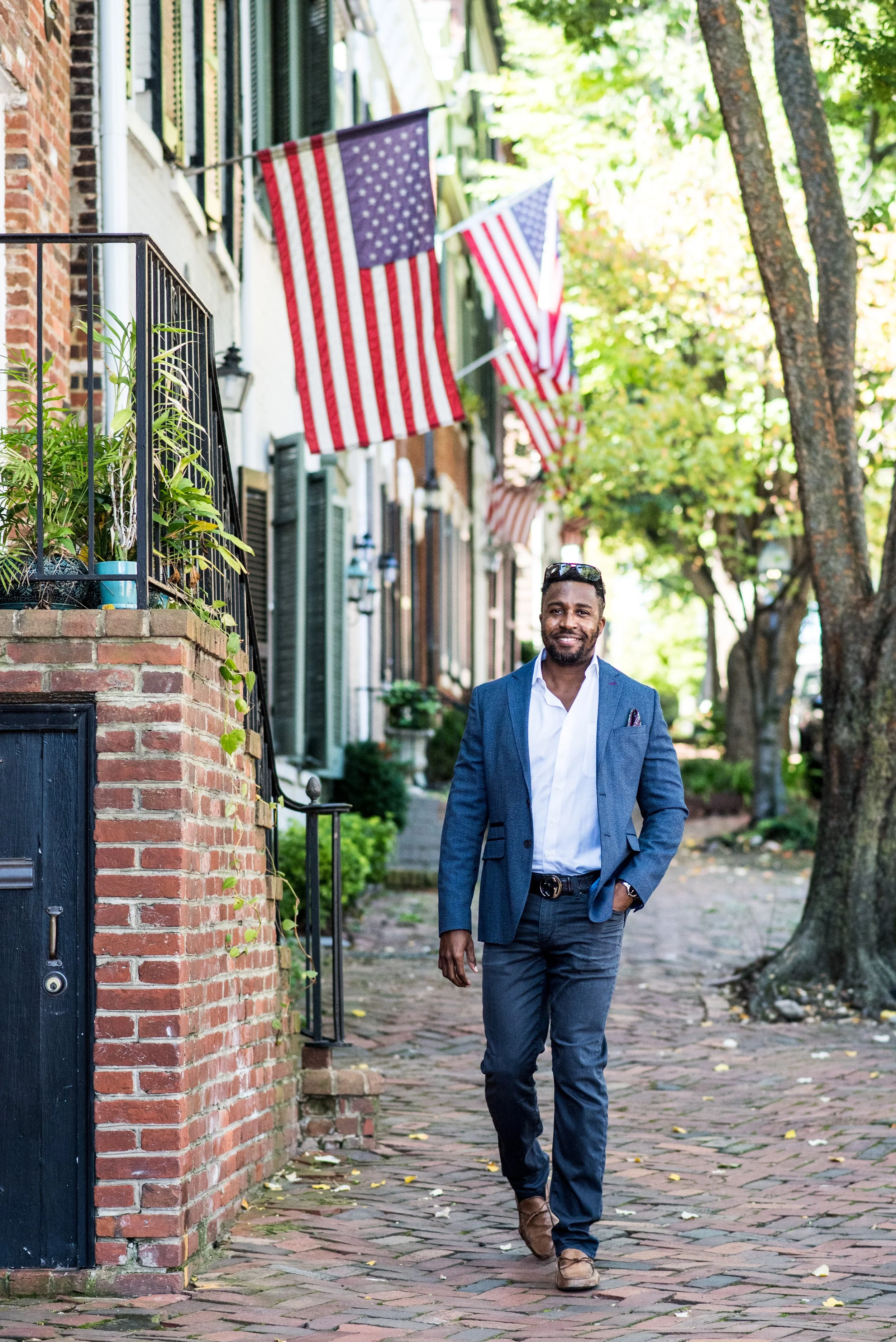 A man in a blue blazer and dress pants walks down a brick sidewalk lined with autumn leaves, with American flags hanging outside a row of houses with green trees in the background.