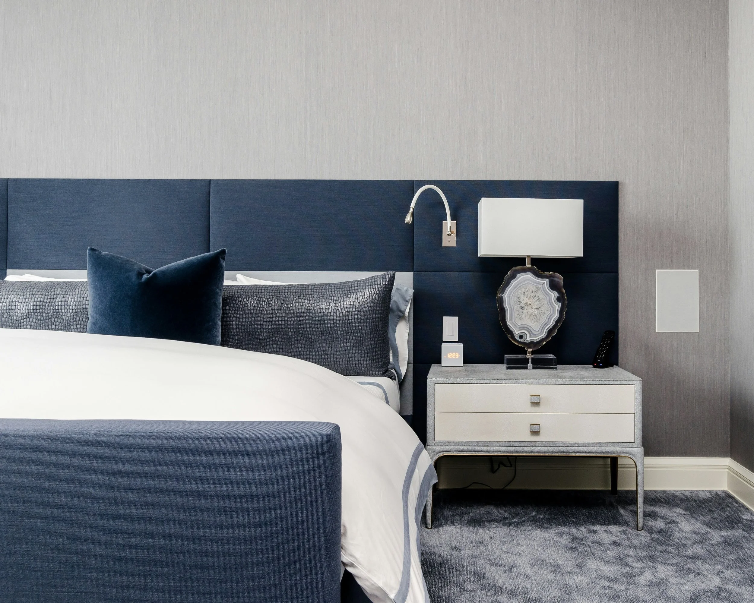 A bedroom with a white bed, dark blue headboard, blue pillows, nightstand with a white lamp, agate slice, digital clock, and a remote control, against a light gray wall and dark carpet.