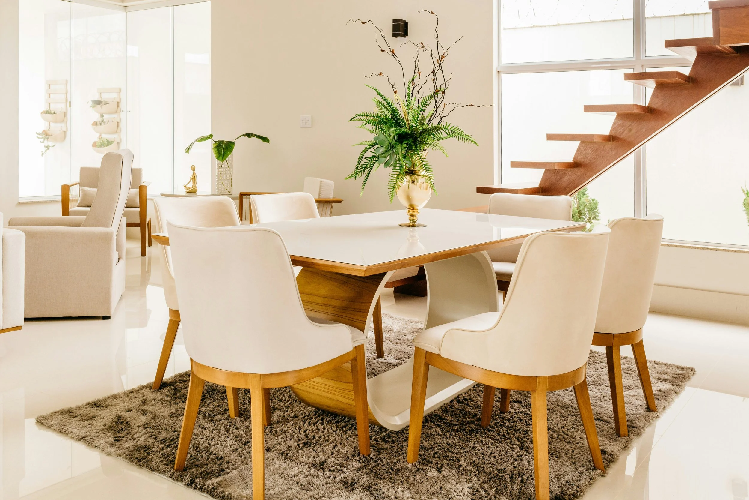 Modern living space with a wooden dining table, white chairs, a large vase with green plants, a beige armchair, a gray rug, large windows, and wooden stairs.