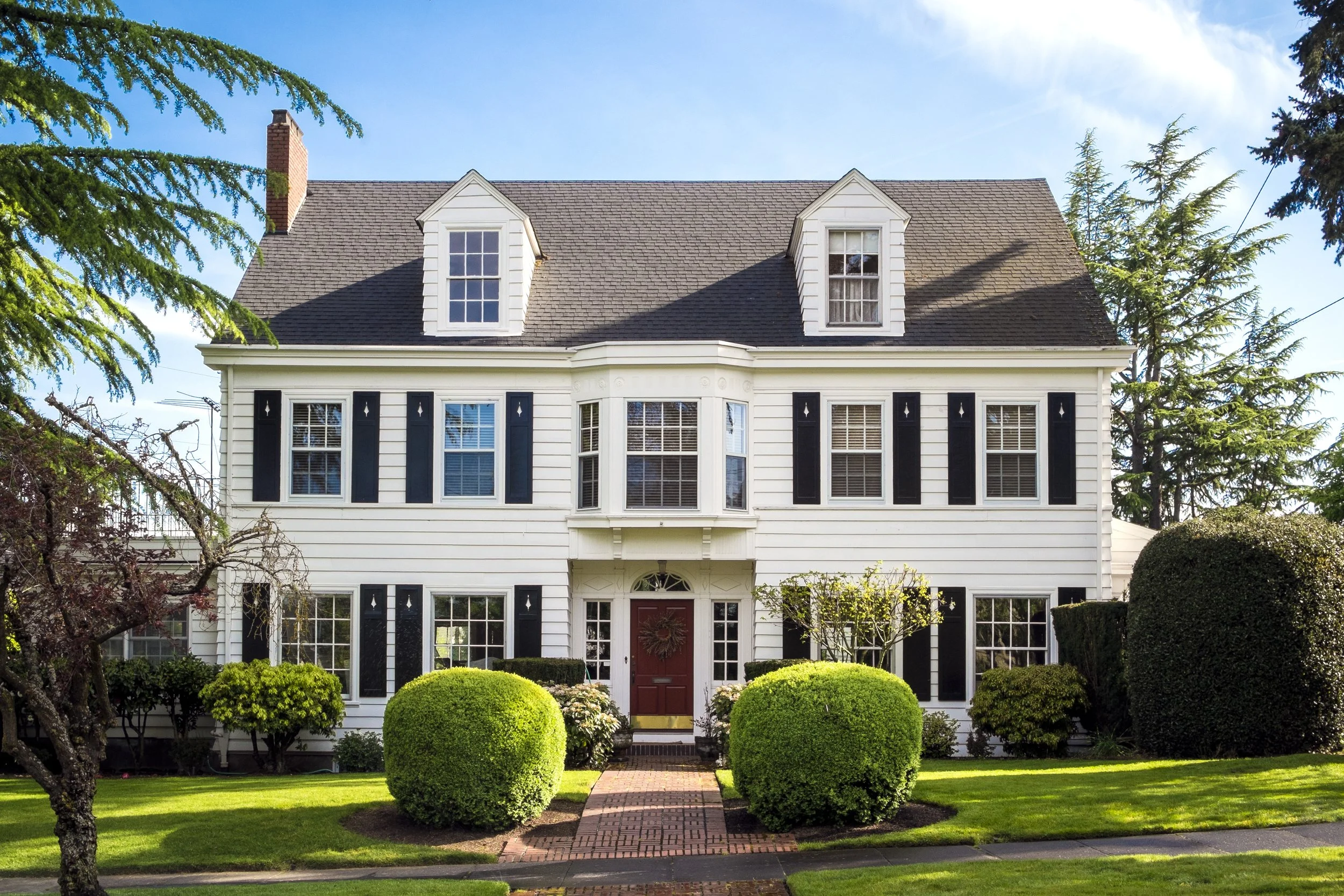 A two-story white house with black shutters and a brick sidewalk leading to the front door, surrounded by a green lawn and manicured bushes, under a blue sky with some clouds.