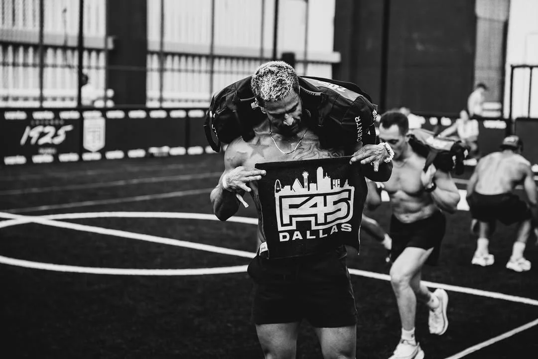 A muscular man in a black tank top and shorts holding a weighted vest labeled 'F45 Dallas' while lifting it in an indoor gym with other men working out in the background.