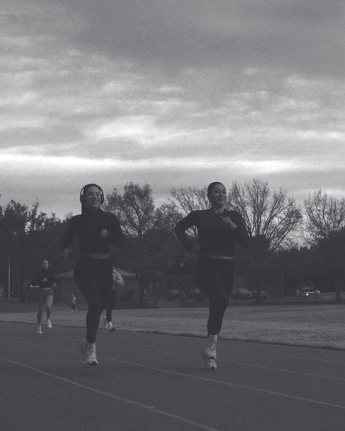 Three women running on a track outdoors during cloudy weather; trees and a building in the background.