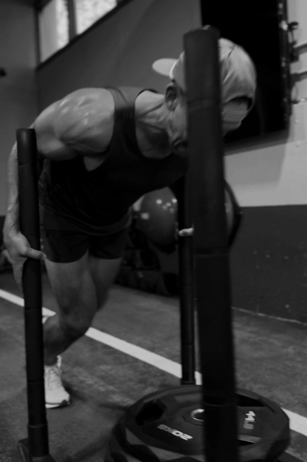 A person in a gym performing a sled push exercise, wearing a cap and athletic clothing.