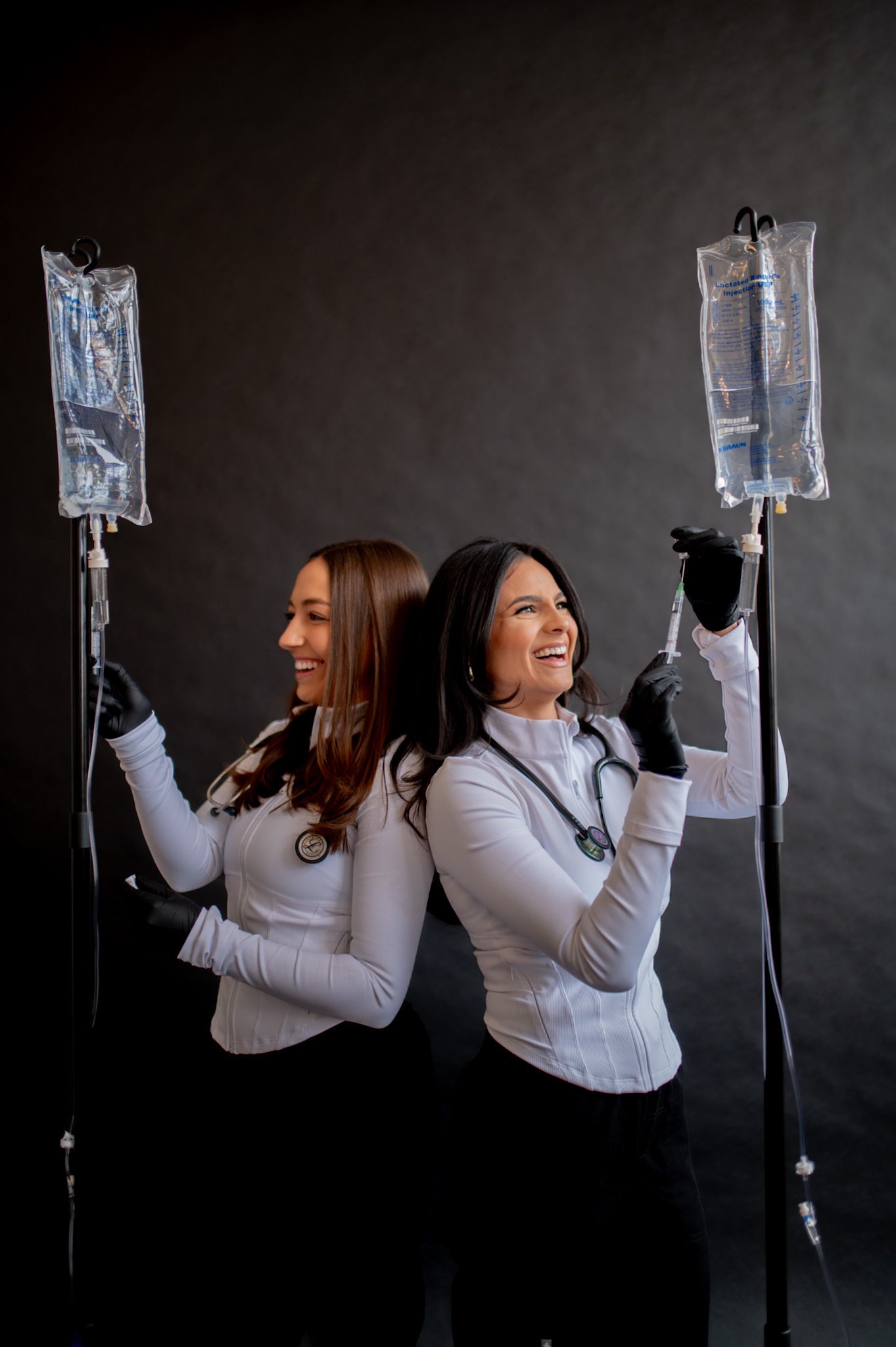 Two female medical professionals in white coats smiling and holding IV bags on stands, standing against a dark background.