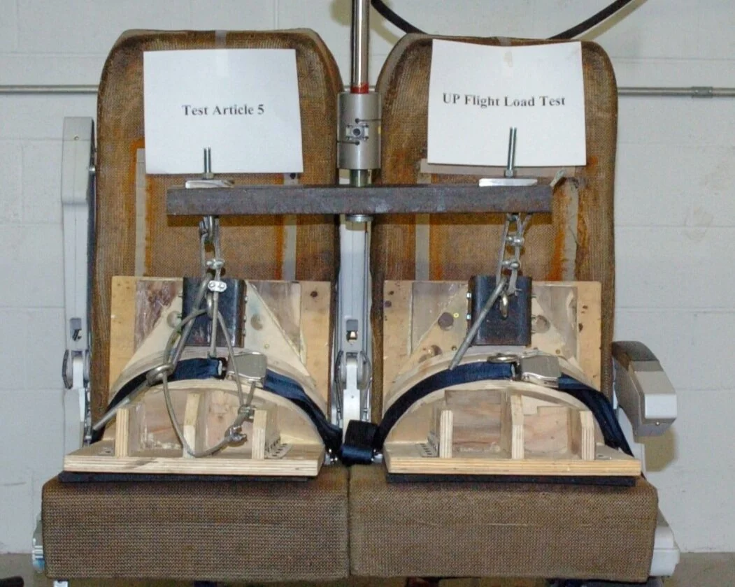 Two aircraft seat structures mounted on a testing fixture with metal brackets, restraint straps, and a central hydraulic actuator for an upward flight load test.