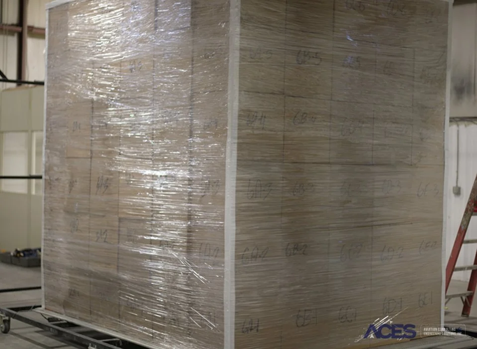 Large rectangular cargo test assembly wrapped in transparent plastic with marked measurement locations, positioned on a metal platform inside the ACES testing laboratory.