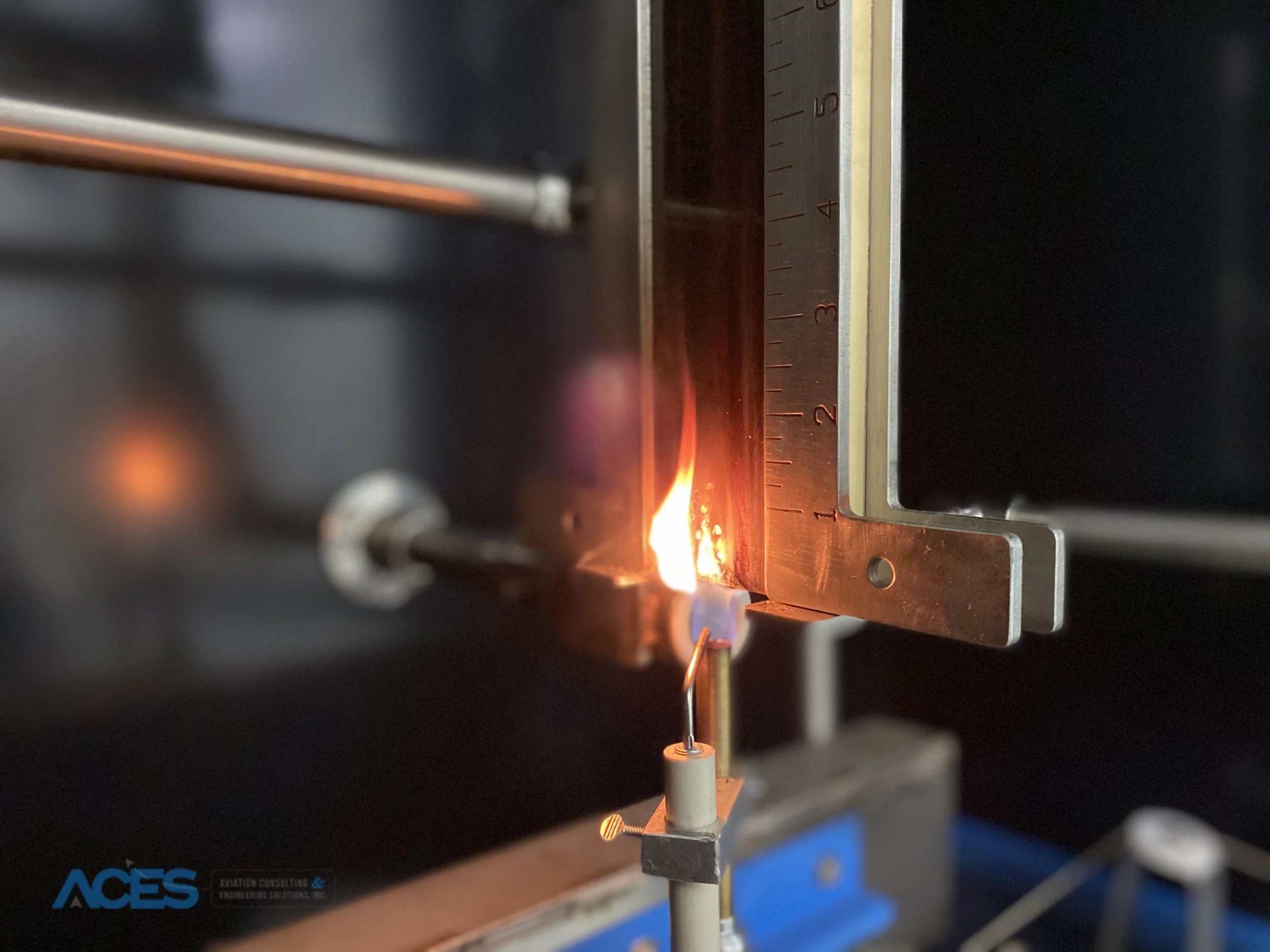 Close-up of a metal specimen holder and brass burner tube inside an aircraft material flammability testing chamber.