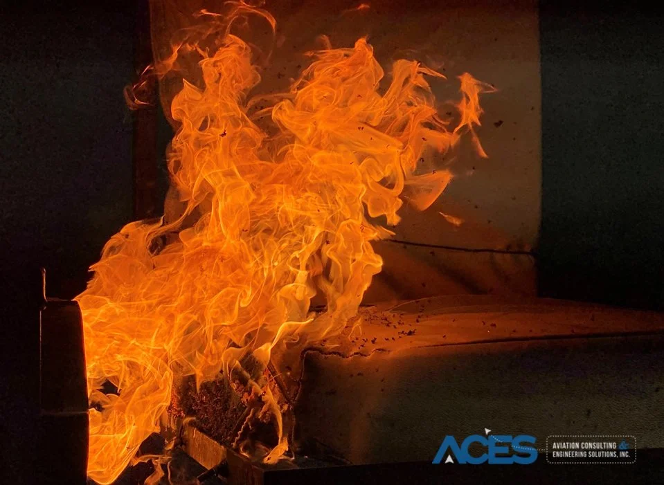 Large orange flames engulfing an aircraft seat cushion test specimen during an oil burner fireblock test in a darkened laboratory.
