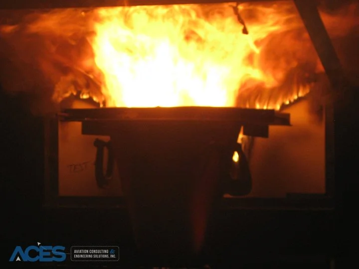 Intense yellow and orange flames engulfing a rectangular cargo liner test assembly inside a darkened fire testing chamber.