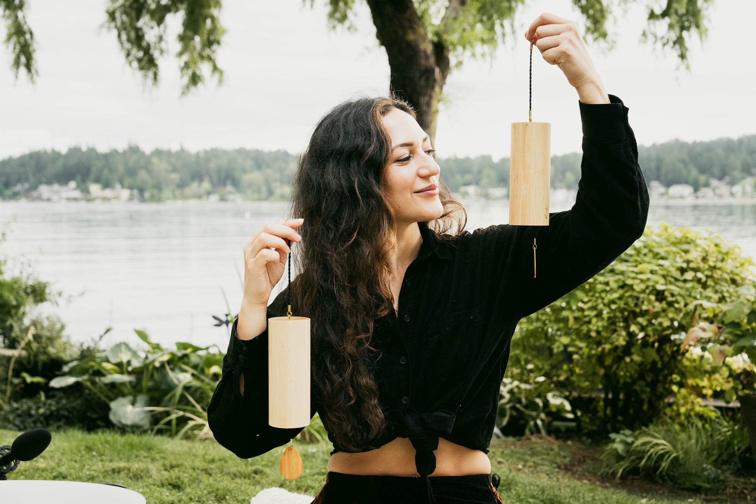 A woman with long, wavy brown hair stands outdoors near a body of water, holding two wooden wind chimes with her arms raised. She is smiling slightly and wearing a black shirt, with greenery and trees in the background.