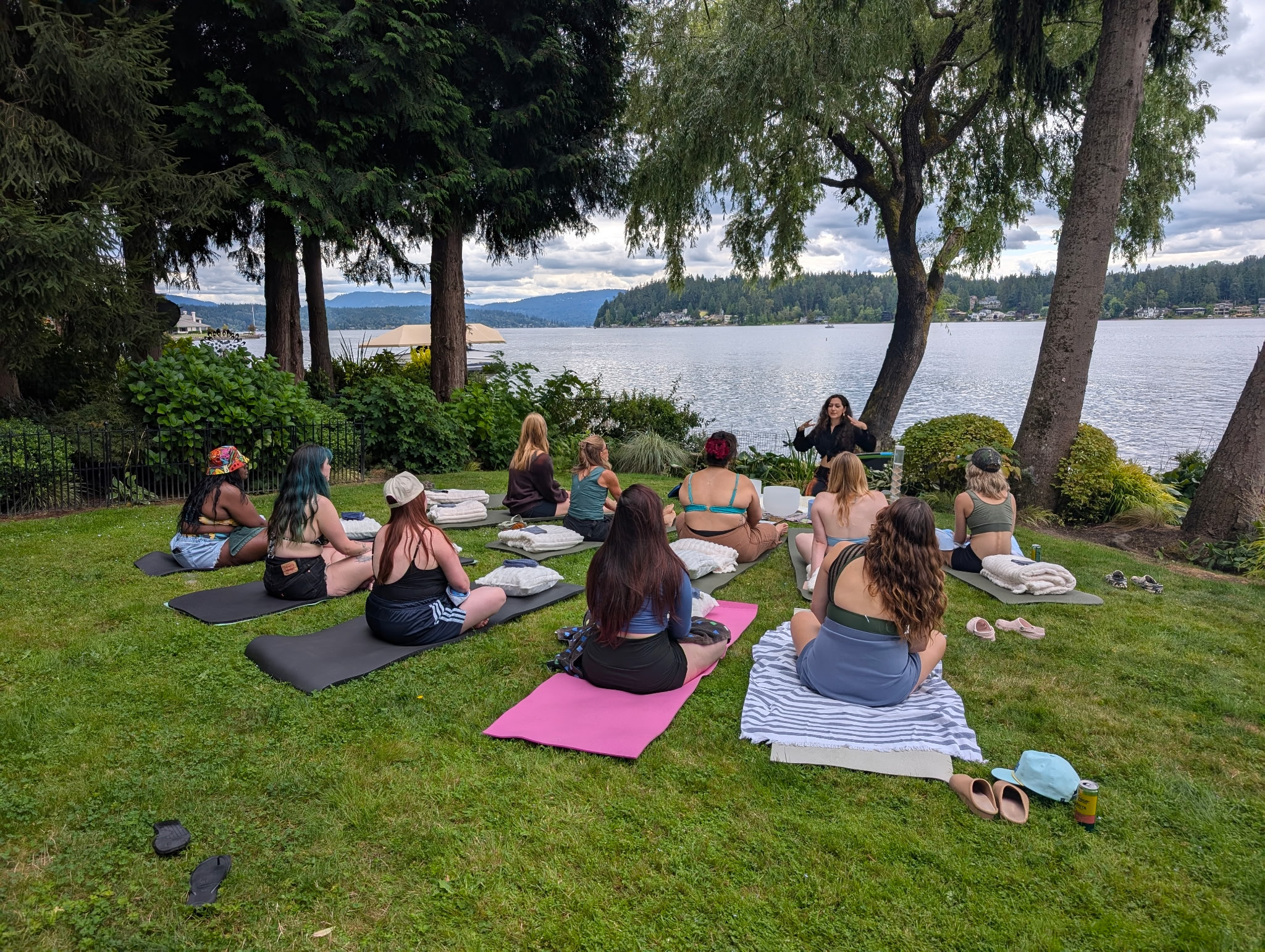 Group of women sitting on yoga mats outdoors near a lake, listening to a woman standing and speaking, with trees and mountains in the background.
