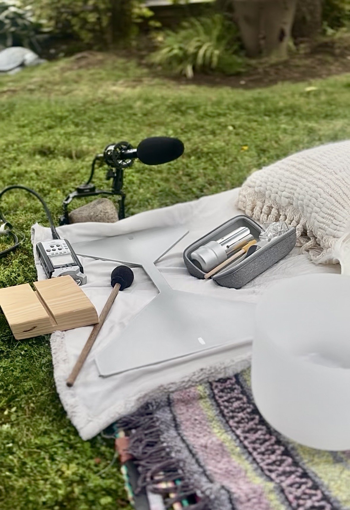 Outdoor recording setup on a blanket includes a microphone with windscreen, a portable audio recorder, a small wooden block, a mallet, a pillow, and a roll of tape, with green grass and trees in the background.