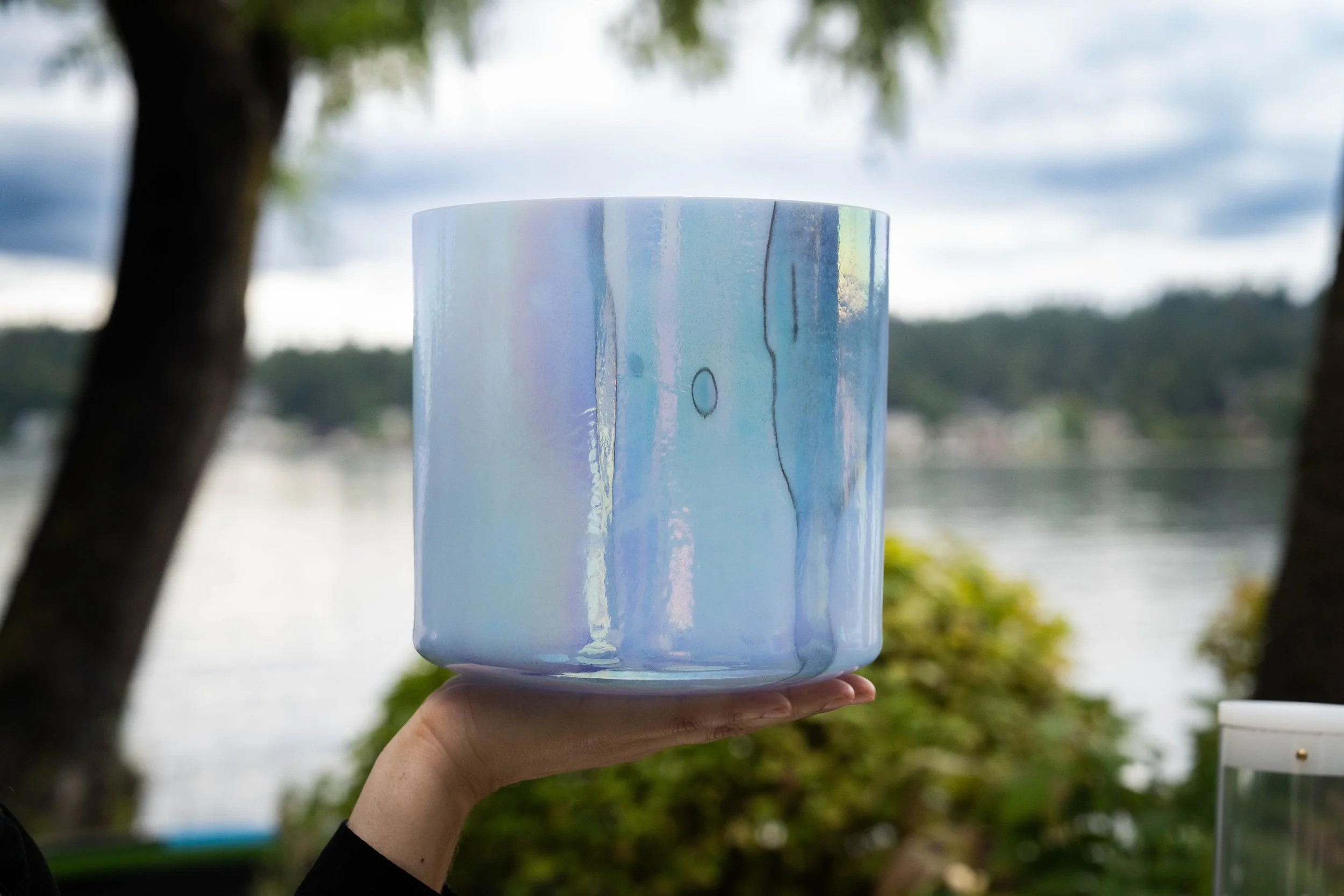 A person holding a ceramic mug with an iridescent glaze outdoors near a body of water, with trees and a cloudy sky in the background.
