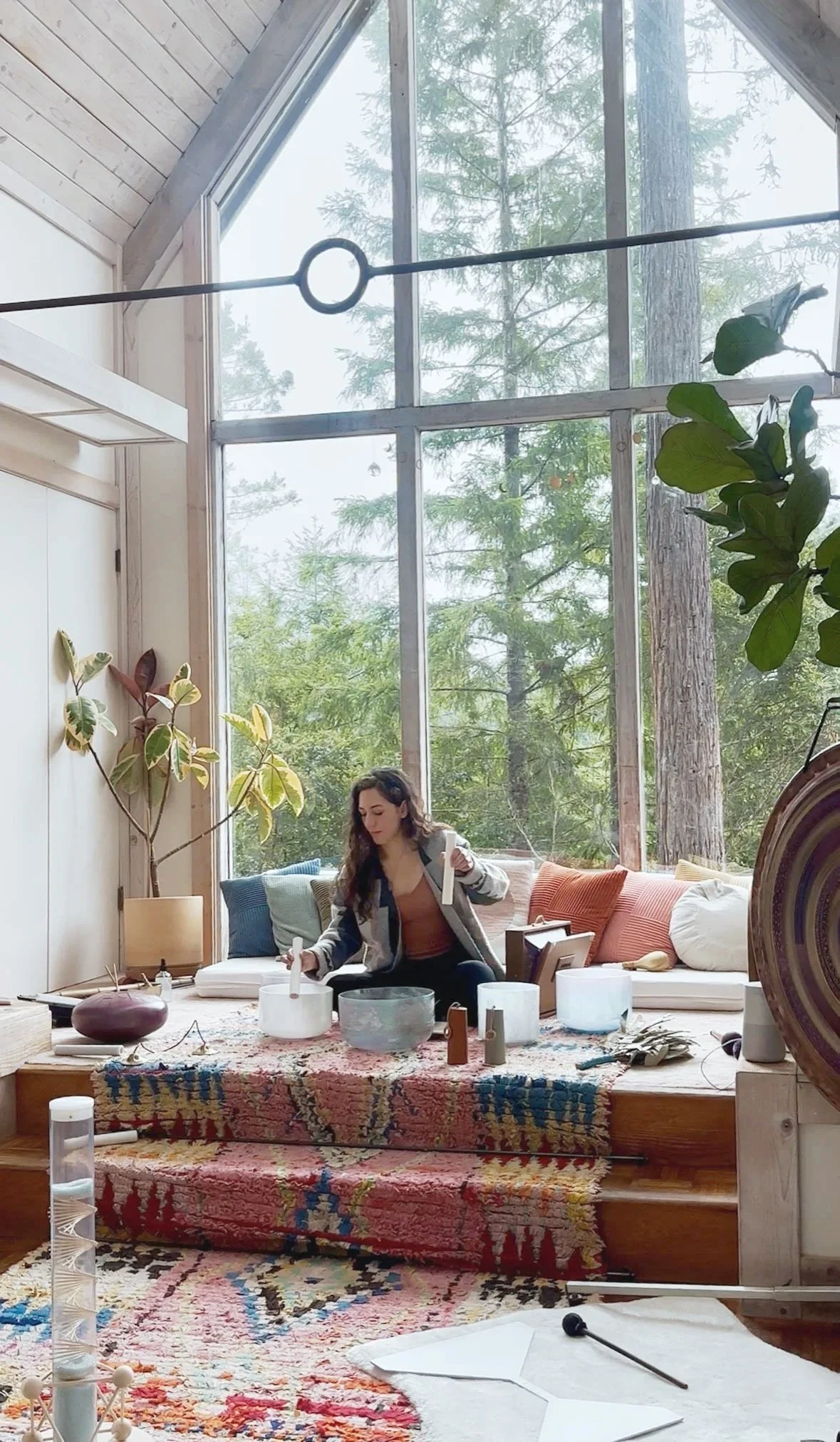Woman sitting on a cushion in a cozy, well-lit room with large windows, surrounded by plants, with various objects and instruments on the floor, engaging with musical or sound instruments.