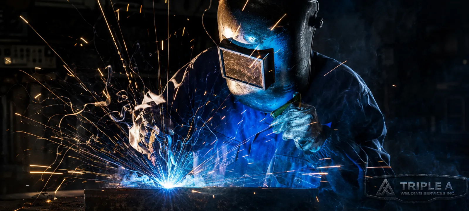 A welder wearing protective gear, including a helmet with a face shield, is welding metal. Sparks and bright blue light are visible as the welding occurs in a dark workshop.