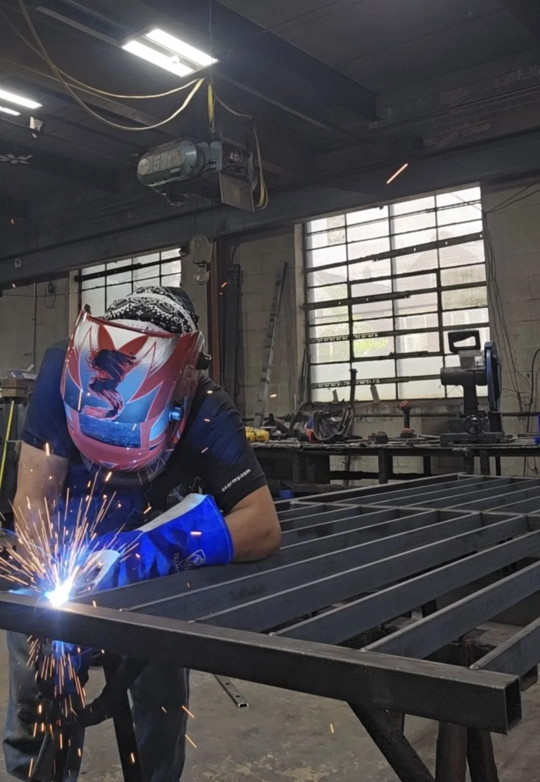 A person wearing a red and black welding helmet and blue gloves welding metal bars in a workshop with tools and equipment around.