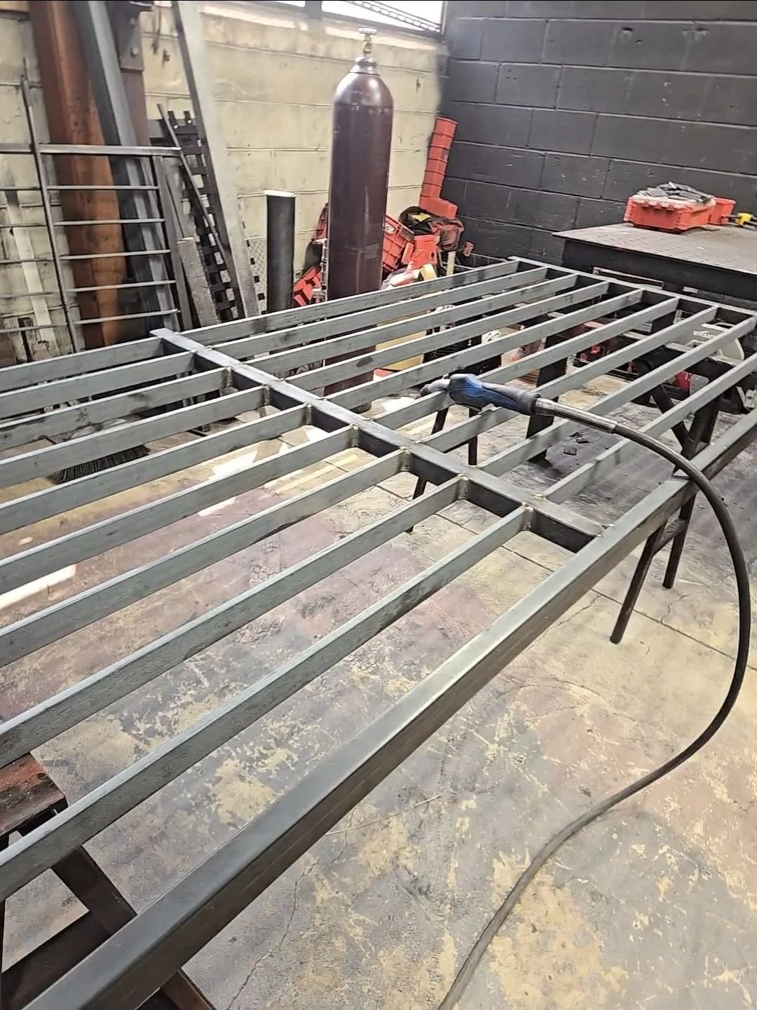 Metal bed frame being welded on a workbench in a workshop with tools and equipment around.