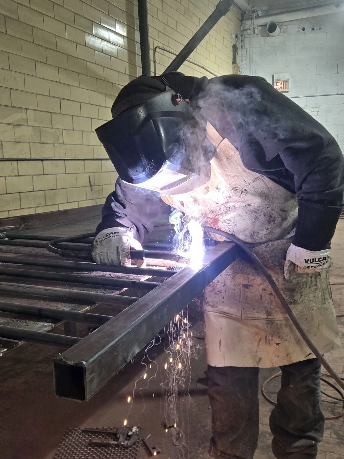 A person wearing a welding helmet and gloves is welding a metal frame, with sparks and smoke visible. The person is leaning over the metal structure in an industrial setting with brick and cinder block walls.