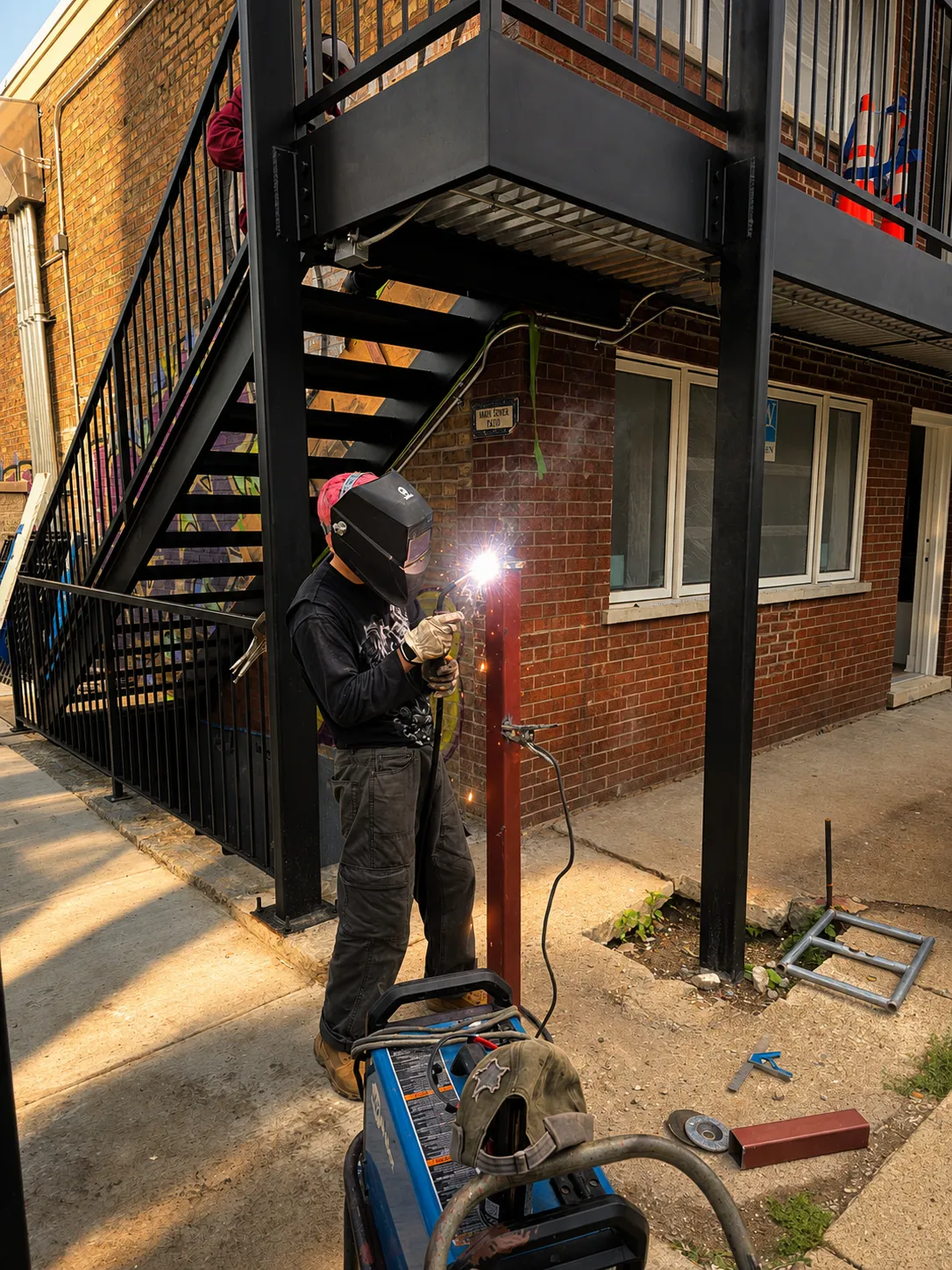Person welding a metal pole on a sidewalk outside a brick building, wearing a protective welding helmet, gloves, and casual clothes, with welding equipment nearby.