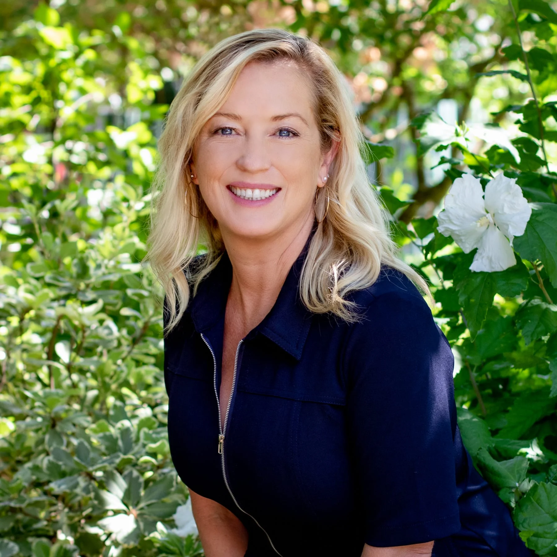 A smiling blonde woman wearing a black zip-up shirt outdoors surrounded by green foliage and white flowers.