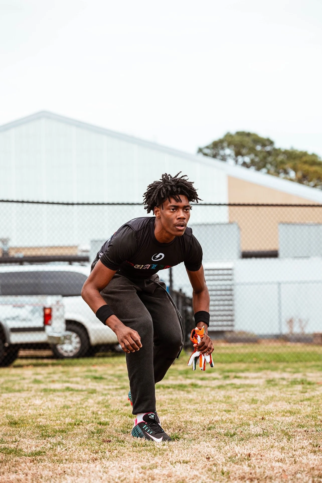 A man practicing football drills on a field, holding gloves in his right hand, wearing black athletic clothes and sneakers, with a chain link fence, vehicles, and a building in the background.