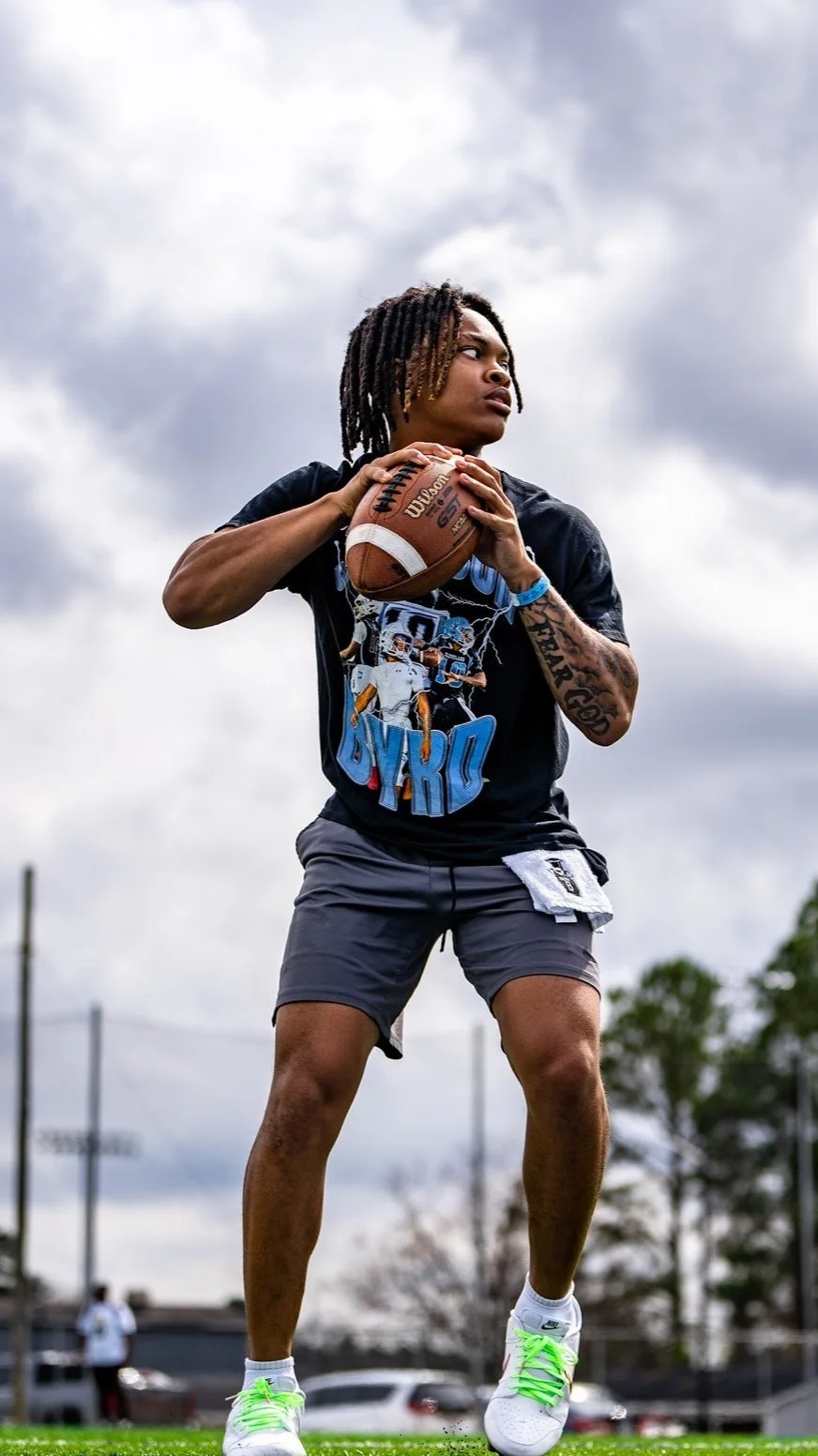 A young man with dreadlocks standing outdoors on a cloudy day, holding a football with both hands, wearing a black T-shirt, gray shorts, white sneakers with neon green laces, and a blue wristband.