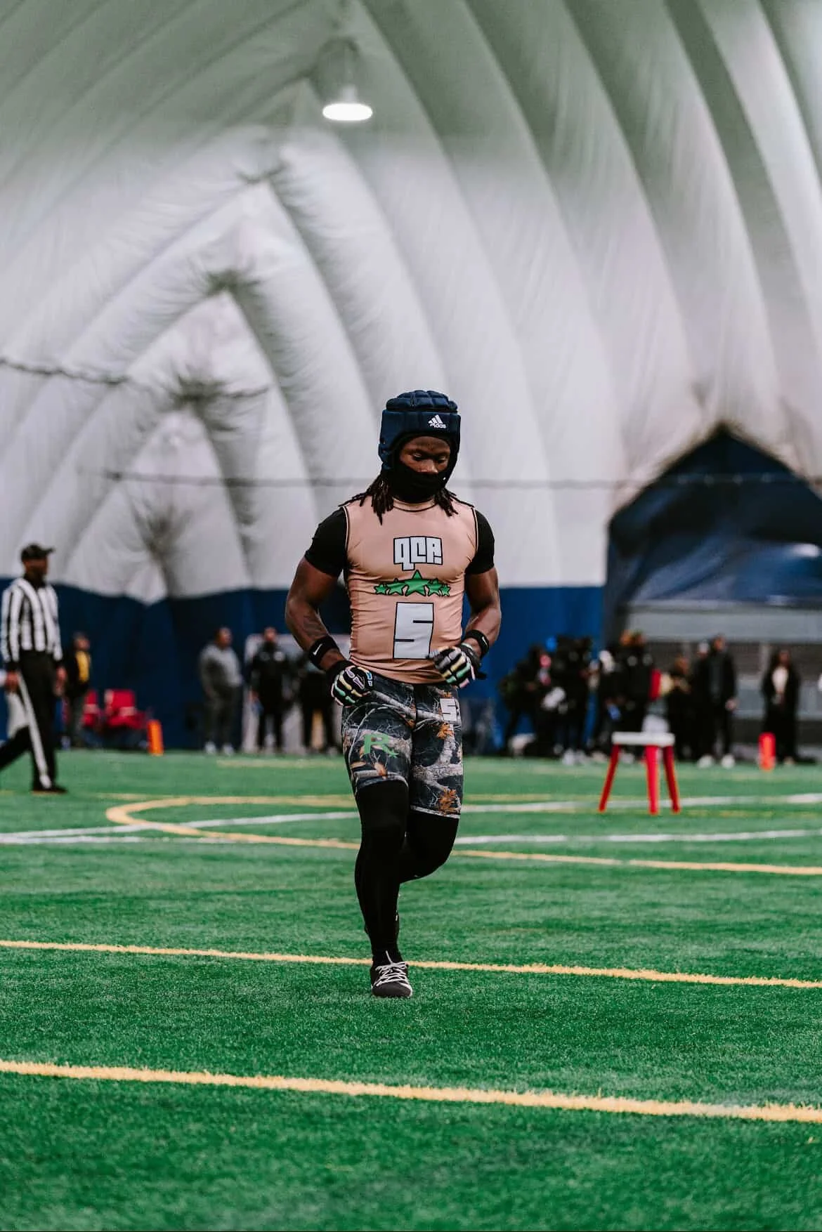 A man in athletic attire walking or jogging on an indoor turf field, with groups of people and a referee in the background inside a large stadium or sports facility.