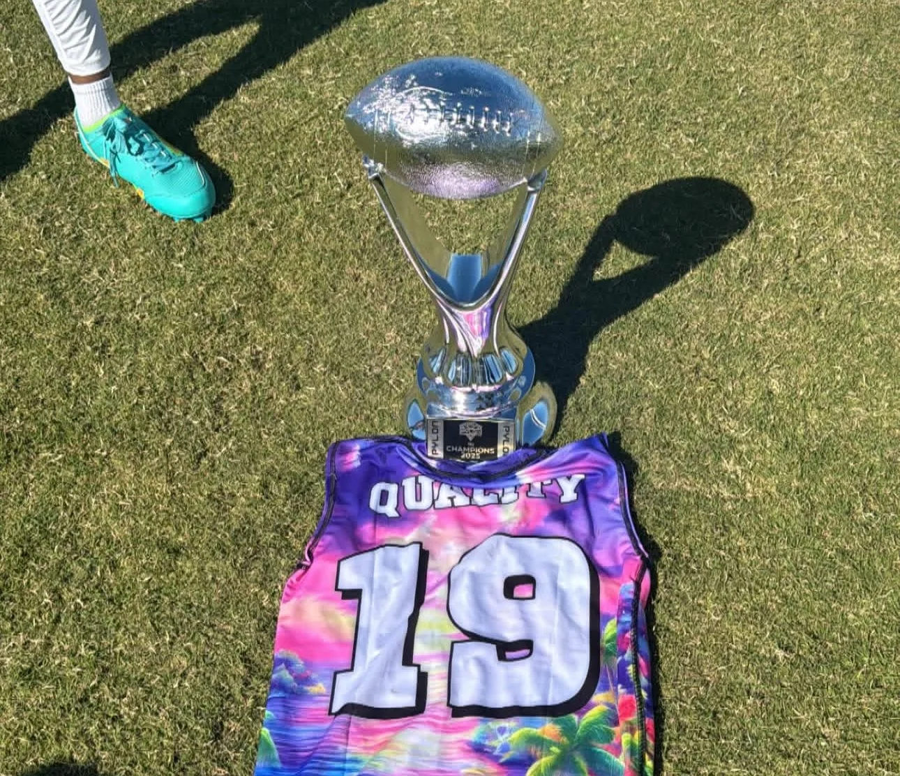 A large silver trophy, a colorful jersey with the number 19, and a black and silver plaque are displayed on a grassy field. Part of a person wearing bright blue cleats and white socks is visible.