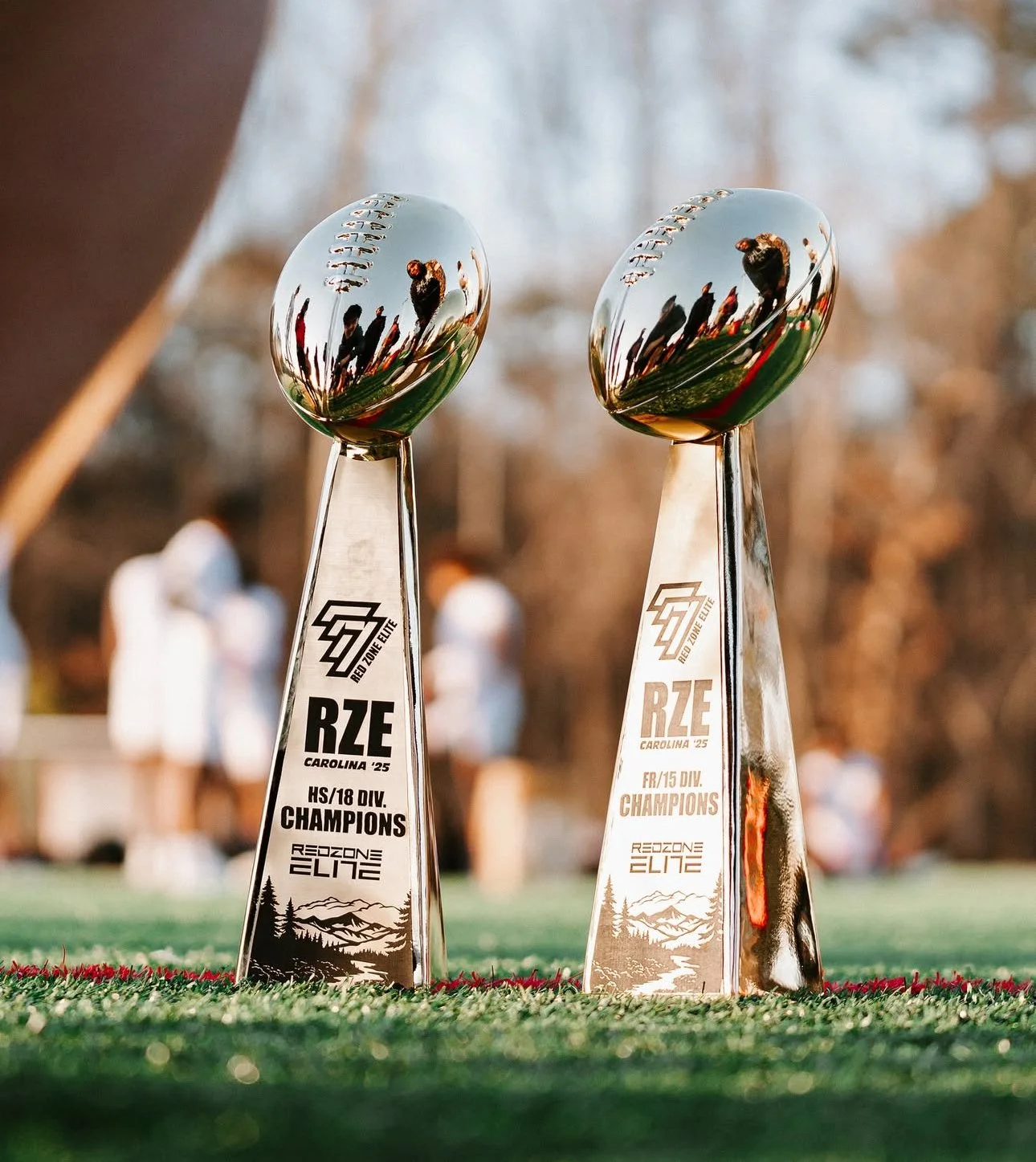 Two silver trophies with reflective, rounded tops and engraved bases displaying Texas-style football trophies. The left trophy's base reads "HS/18 DIV. CHAMPIONS" and the right reads "FR/15 DIV. CHAMPIONS." Both trophies are from the Red Zone Elite e