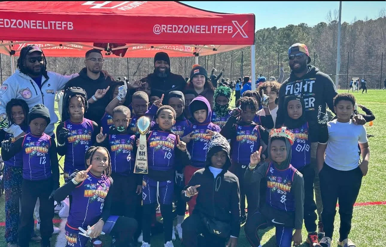 A group of children in purple sports uniforms holding a trophy and making victory signs, surrounded by adults at a sporting event on a grassy field with a red tent in the background.