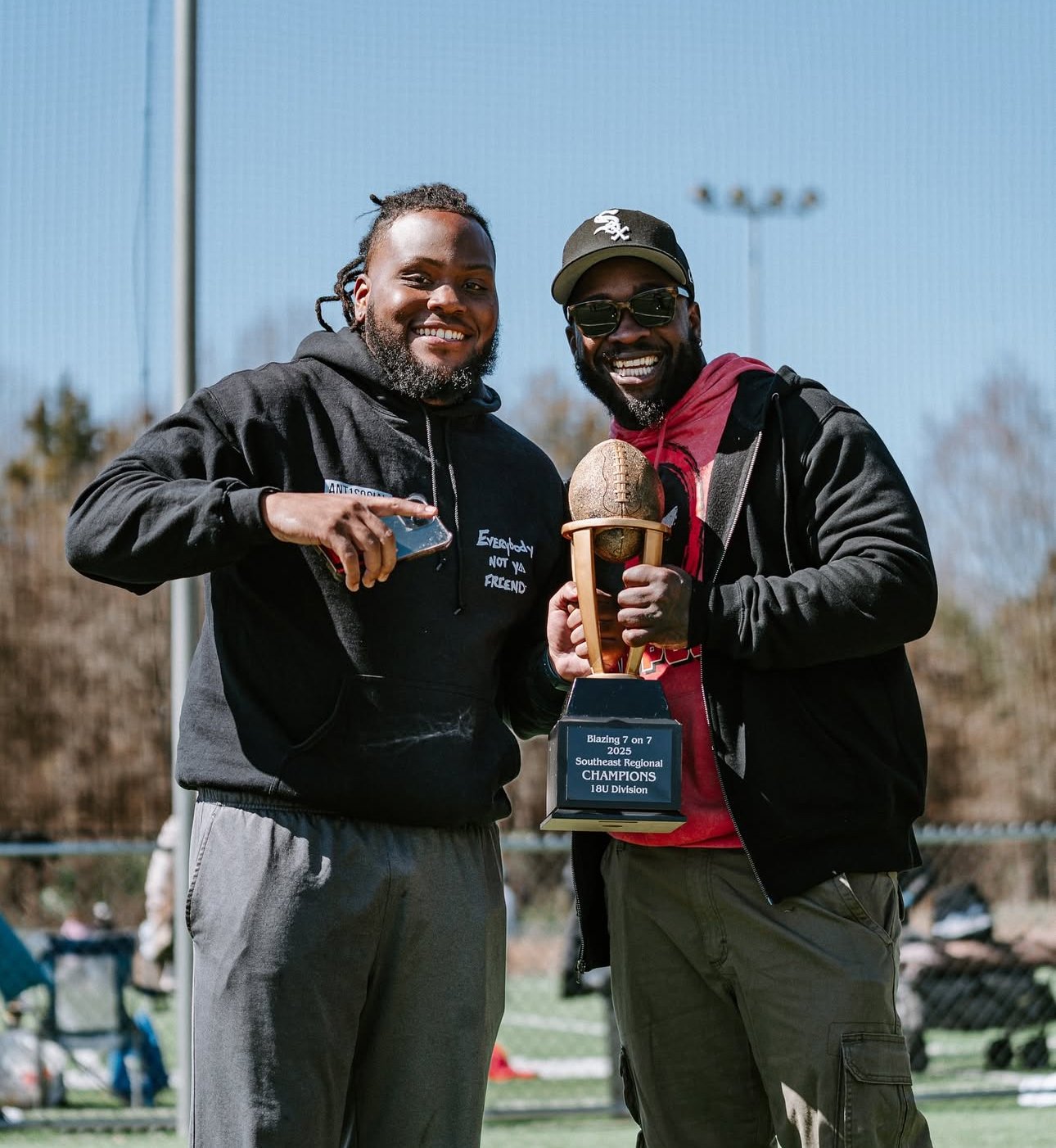 Two men smiling and holding a trophy shaped like a football on a wooden stand, at an outdoor sports field on a sunny day.
