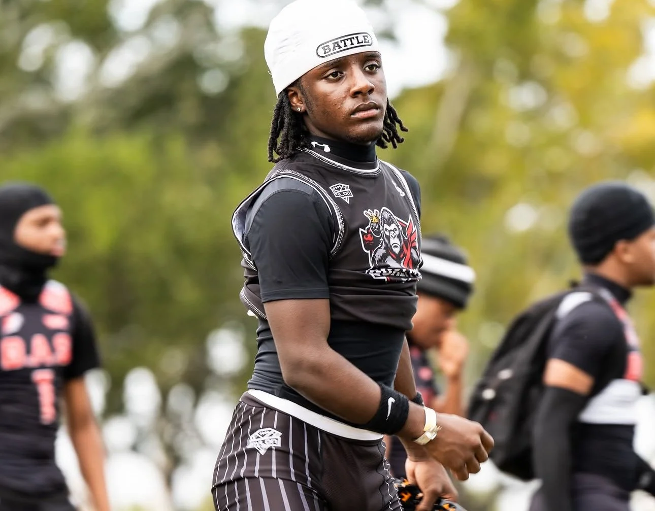 A young man wearing a white head covering and black athletic shirt with a lion graphic. He is outdoors among other people also dressed in black. The background has blurred trees.