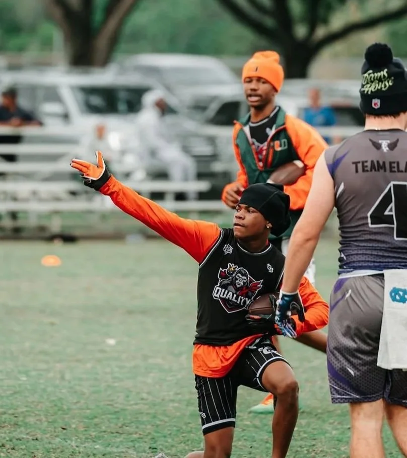 Young football player in a black beanie and orange jacket with black and white striped shorts reaching out during a game, with a coach or teammate in gray and purple uniform nearby, and another person in an orange beanie in the background.