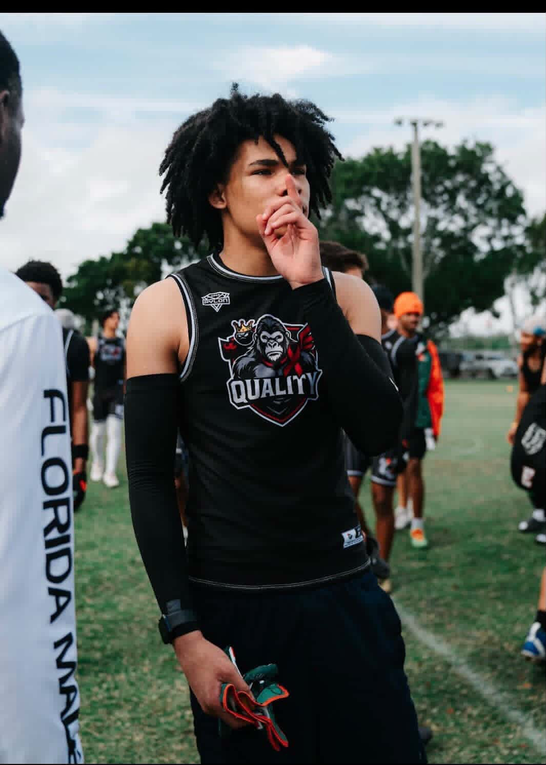 A woman with curly black hair wearing a black sports top with a logo featuring a gorilla and the word 'QUALITY,' standing on a grassy field among other athletes during an outdoor sports event.