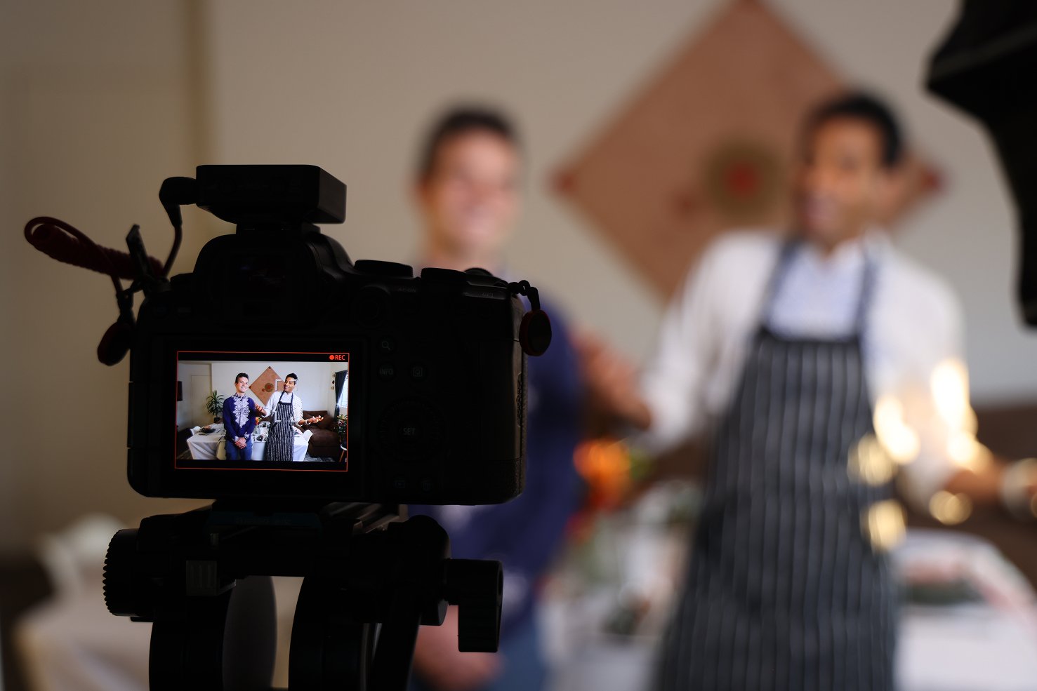 A camera recording two people, one in a suit and one in a chef's uniform, during a filming or interview in an indoor setting.