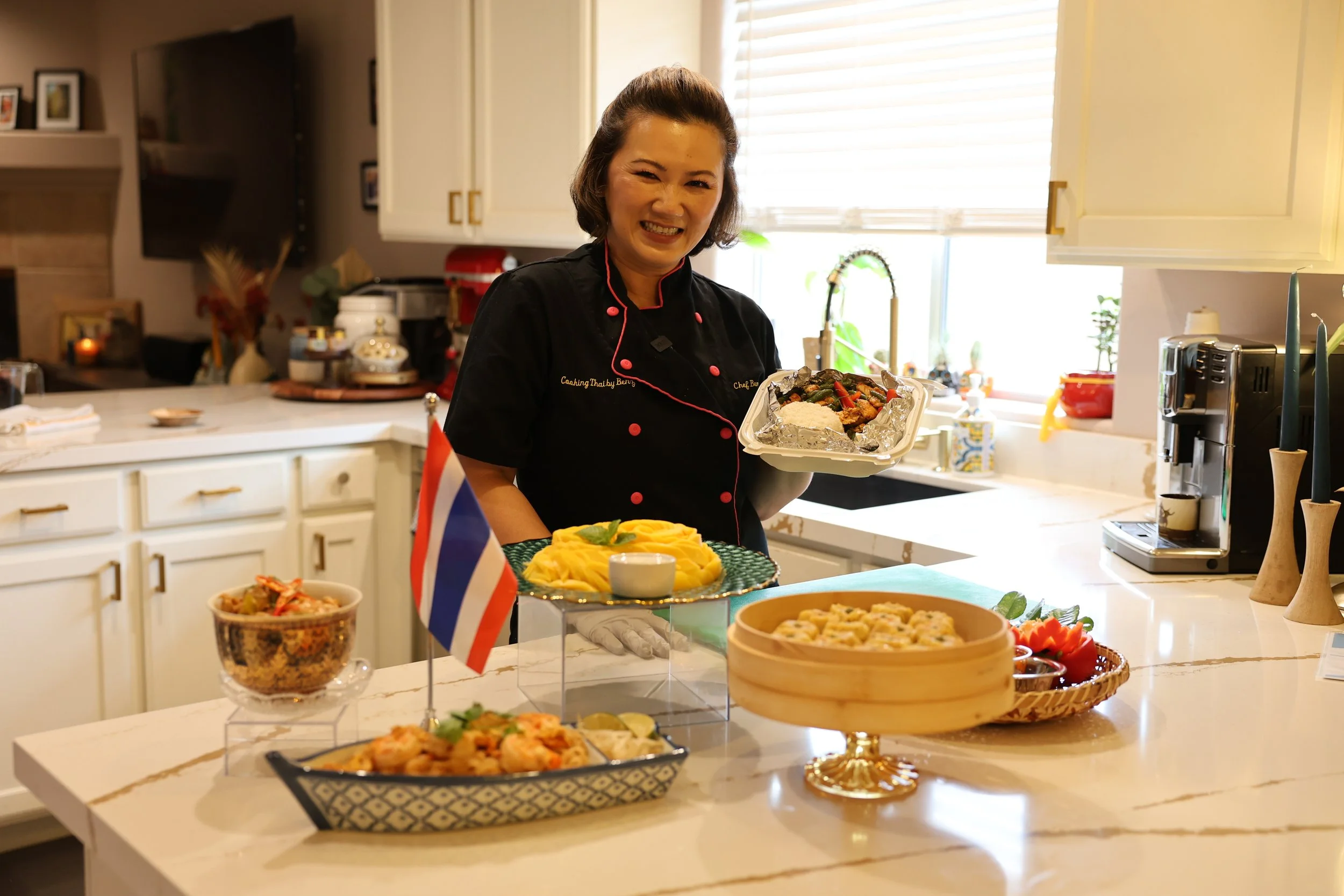 A woman in a black chef's uniform smiling and holding a dish of food in a bright kitchen. In the foreground, there are various dishes and decorations on the counter, including a small flag. The kitchen has white cabinets, a window with blinds, and kitchen appliances.