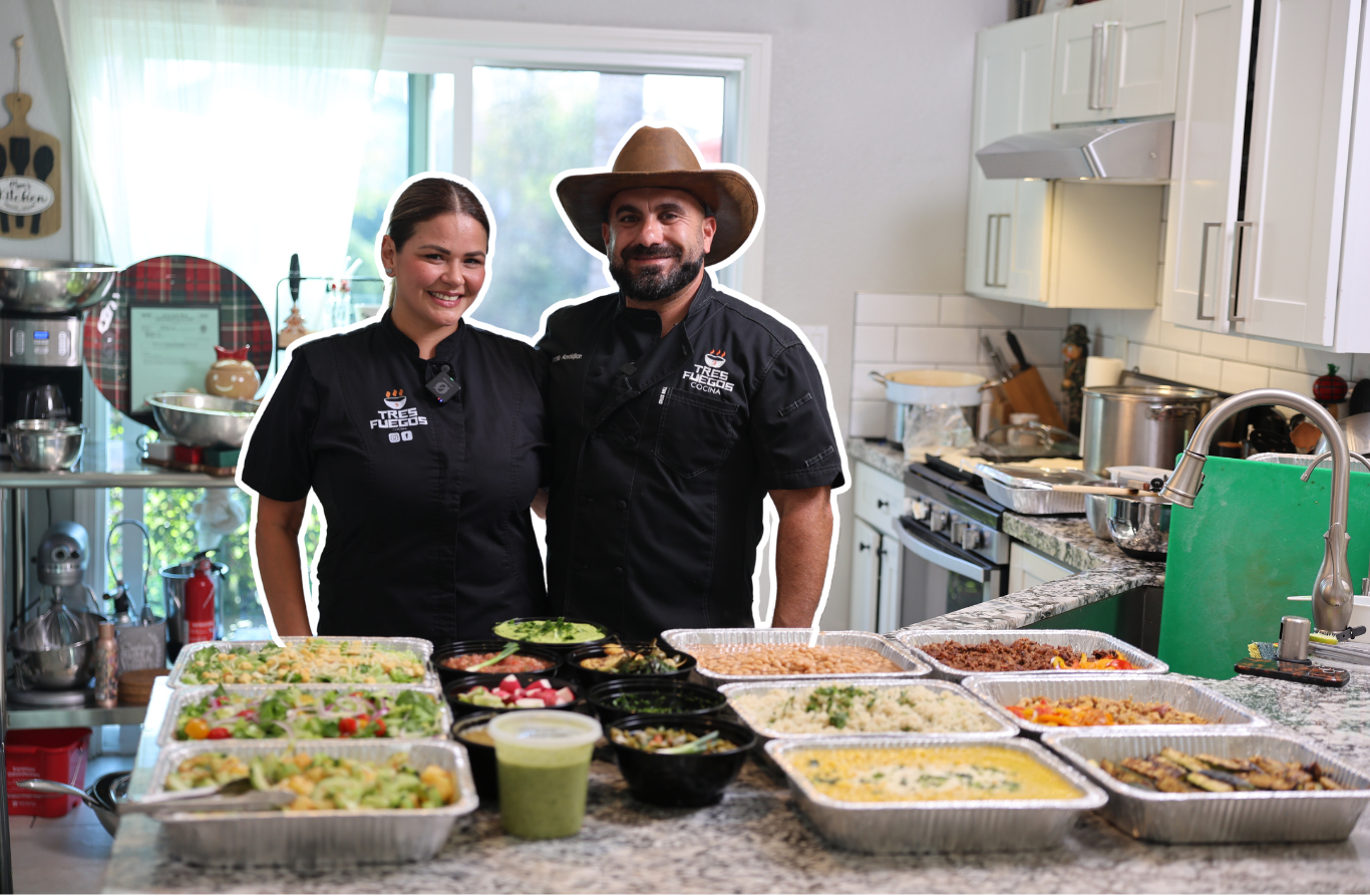 Two MEHKO chefs standing in a kitchen surrounded by various prepared dishes on the counter.