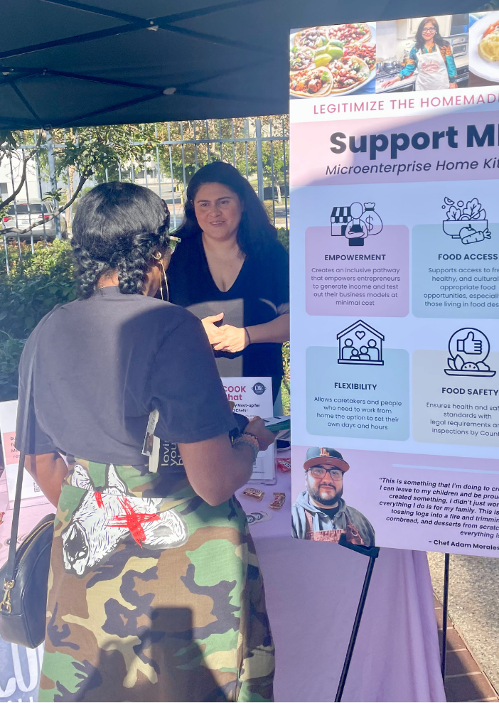 Two women standing at an outdoor information booth. One woman is wearing glasses and a black shirt, engaging with the other woman who has gray hair, glasses, and is dressed in camouflage pants with a cartoon skull patch, and a black top. There is a large pink and white informational poster about supporting microenterprise home kitchens, listing benefits like empowerment, food access, flexibility, and food safety.