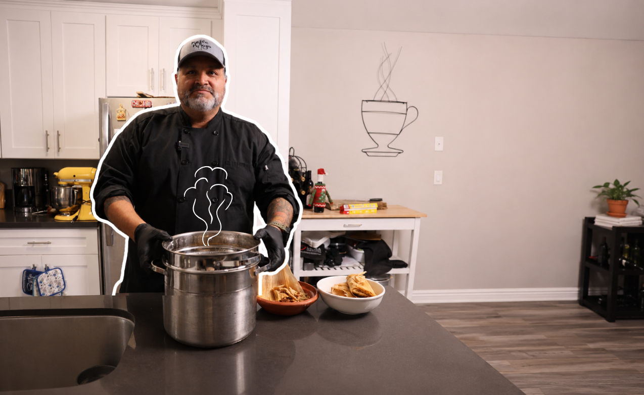 A man in a black chef's coat and a white cap standing in a modern kitchen, holding a steaming pot, with bowls of cooked food on the counter in front of him, and a drawing of a steaming cup on the wall behind him.