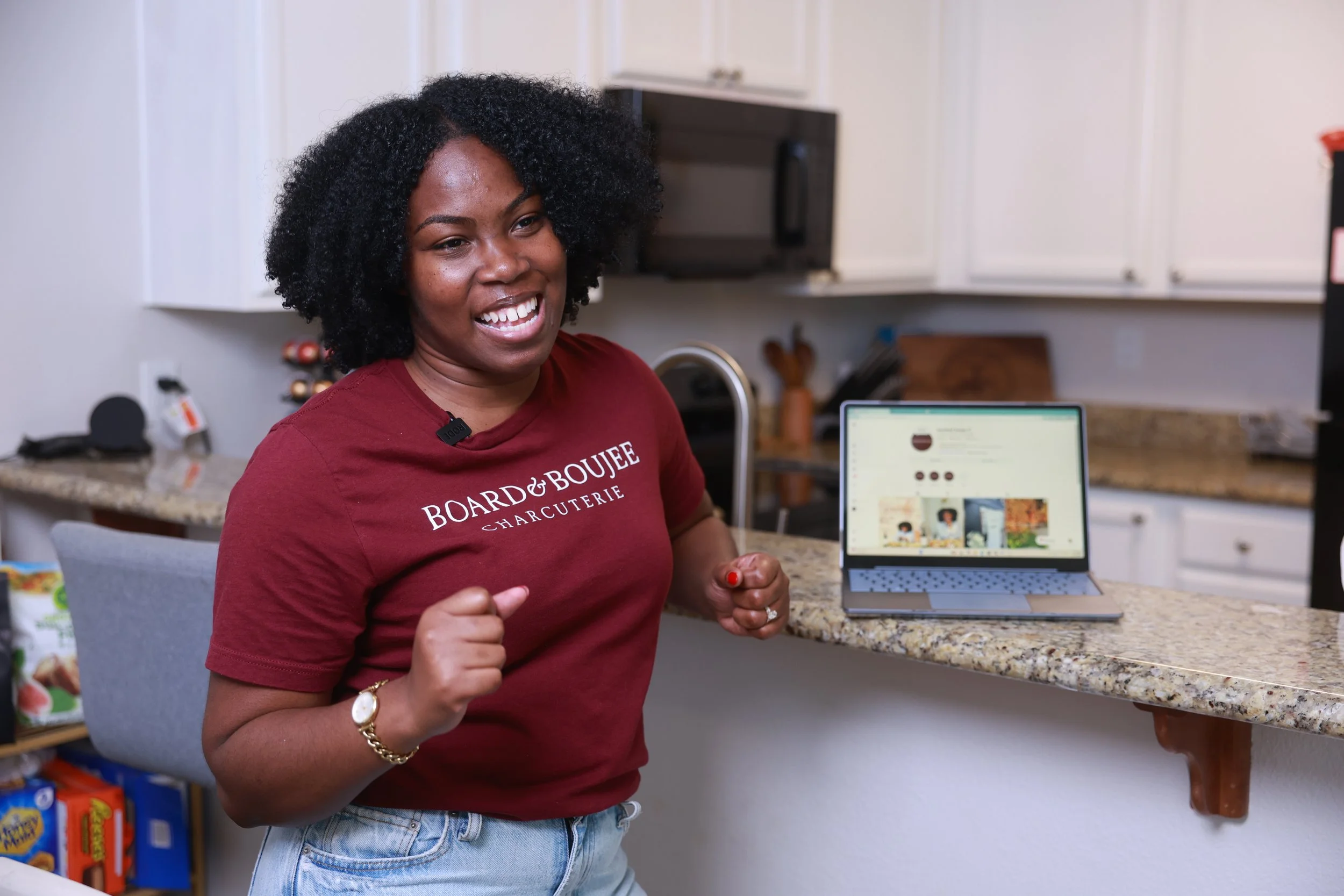 A woman with curly black hair smiling and wearing a red t-shirt with white text, standing in a kitchen beside a laptop on a granite countertop discussing her MEHKO business.