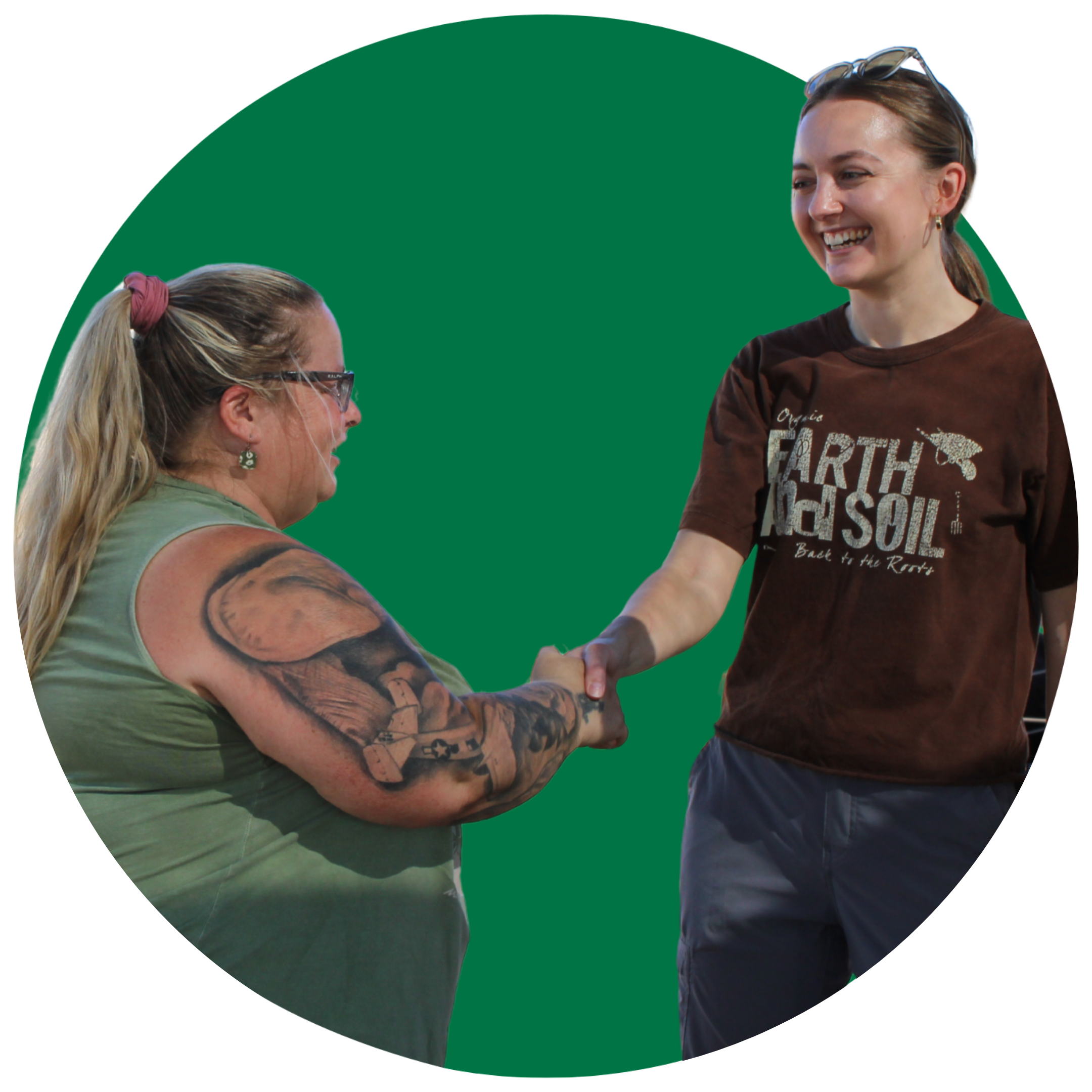 Two women shake hands and smile at each other against a green background.
