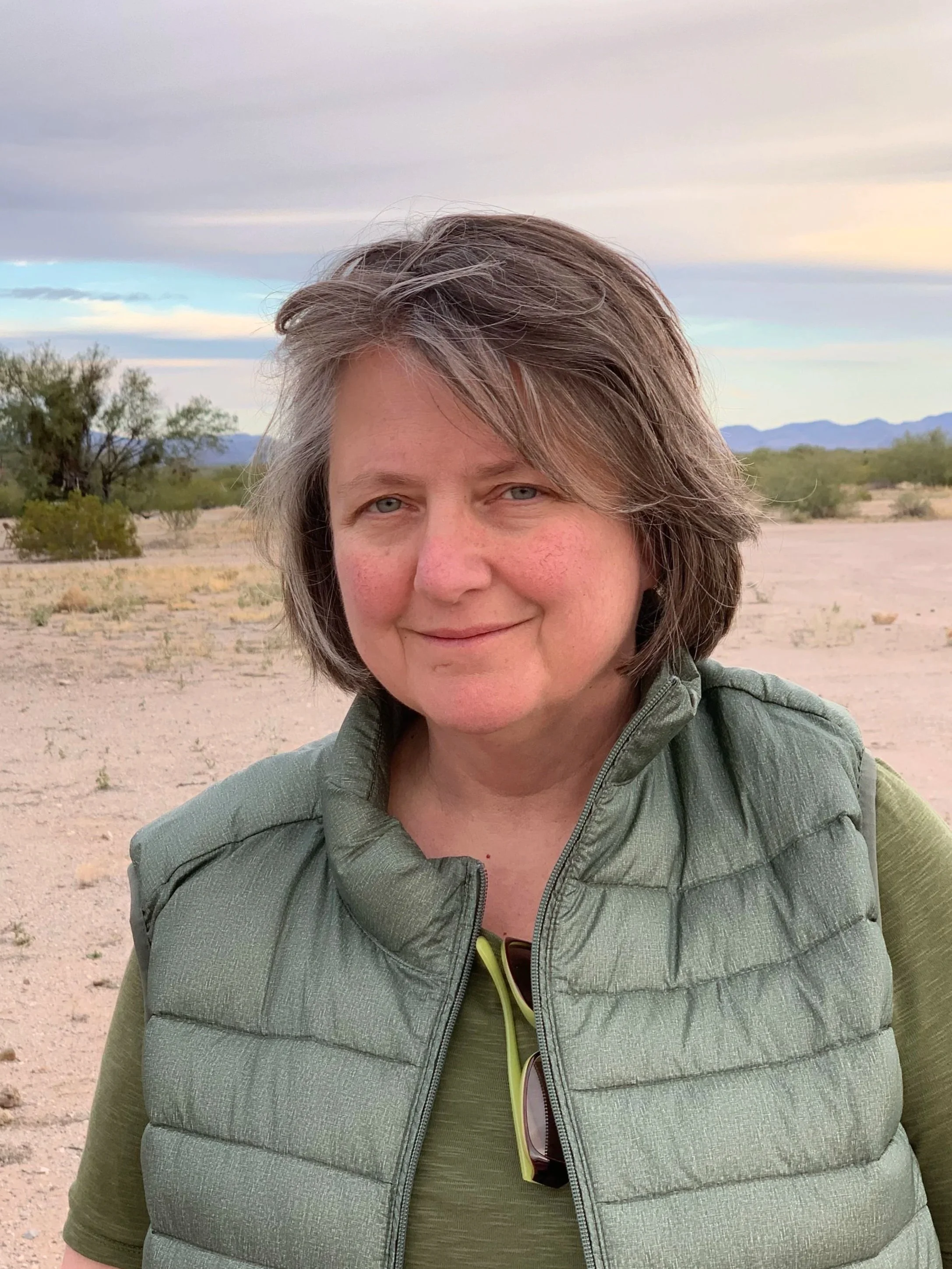 Close-up of Kathleen Merrigan. She has short brown hair and blue eyes, wearing a green shirt and a gray puffer vest, standing outdoors in a desert landscape with mountains, sparse vegetation, and a cloudy sky.
