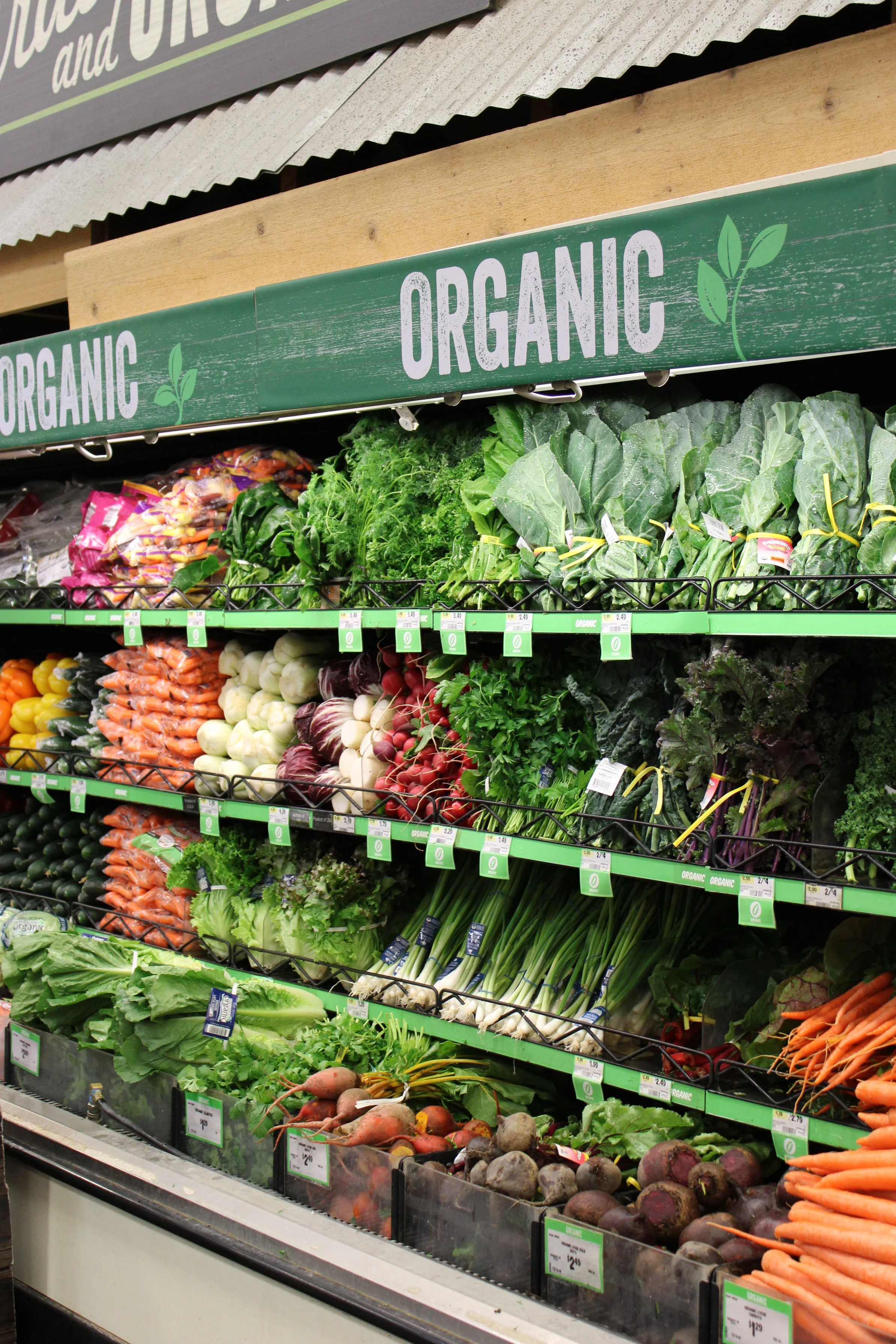 Fresh organic vegetables including carrots, radishes, beets, leafy greens, and celery arranged on grocery store shelves.