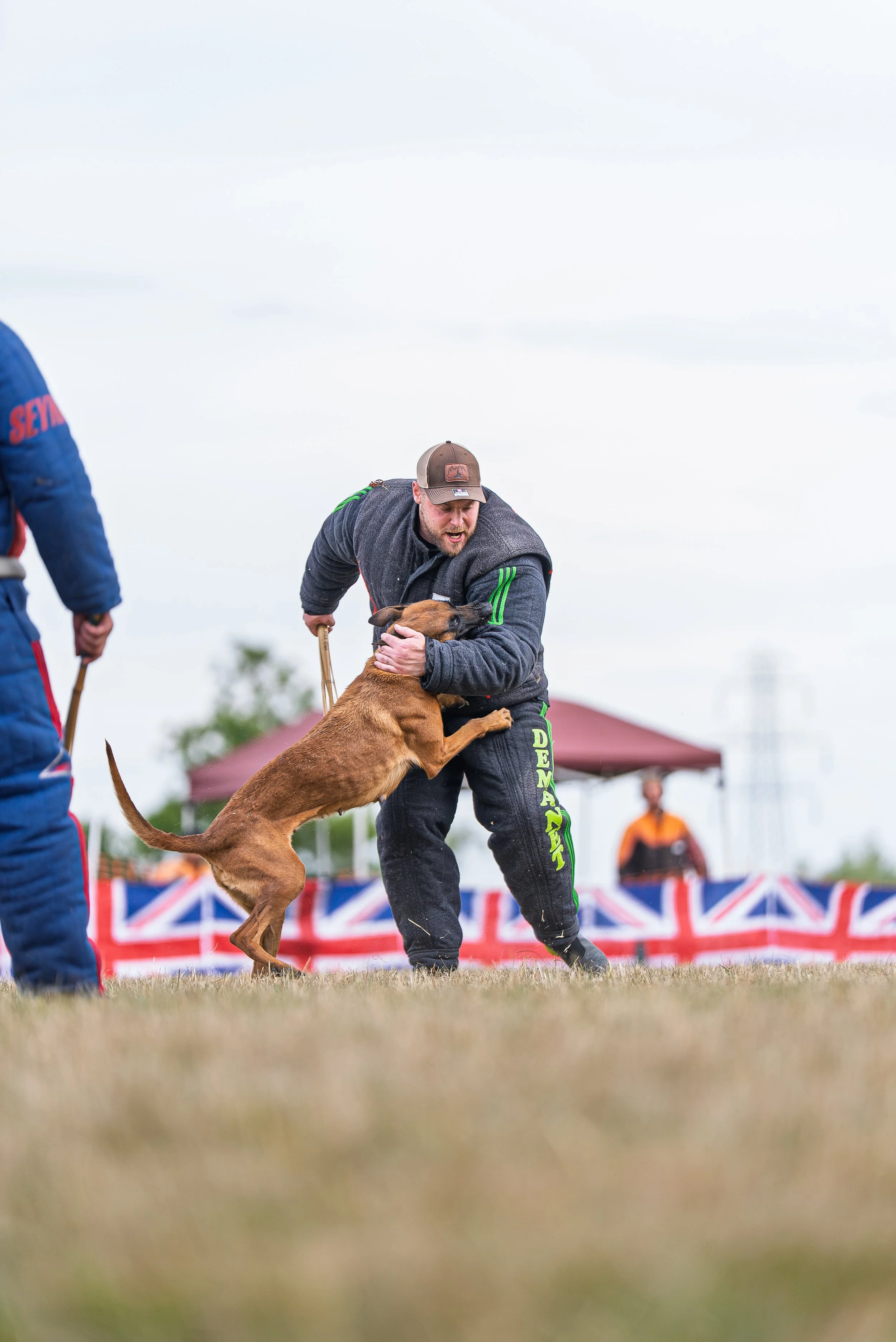 A man in athletic clothing and a cap is holding a dog back during a dog agility competition. The dog, a large brown breed, is jumping and pulling on the man's arm, and there are other participants and a Union Jack banner in the background.