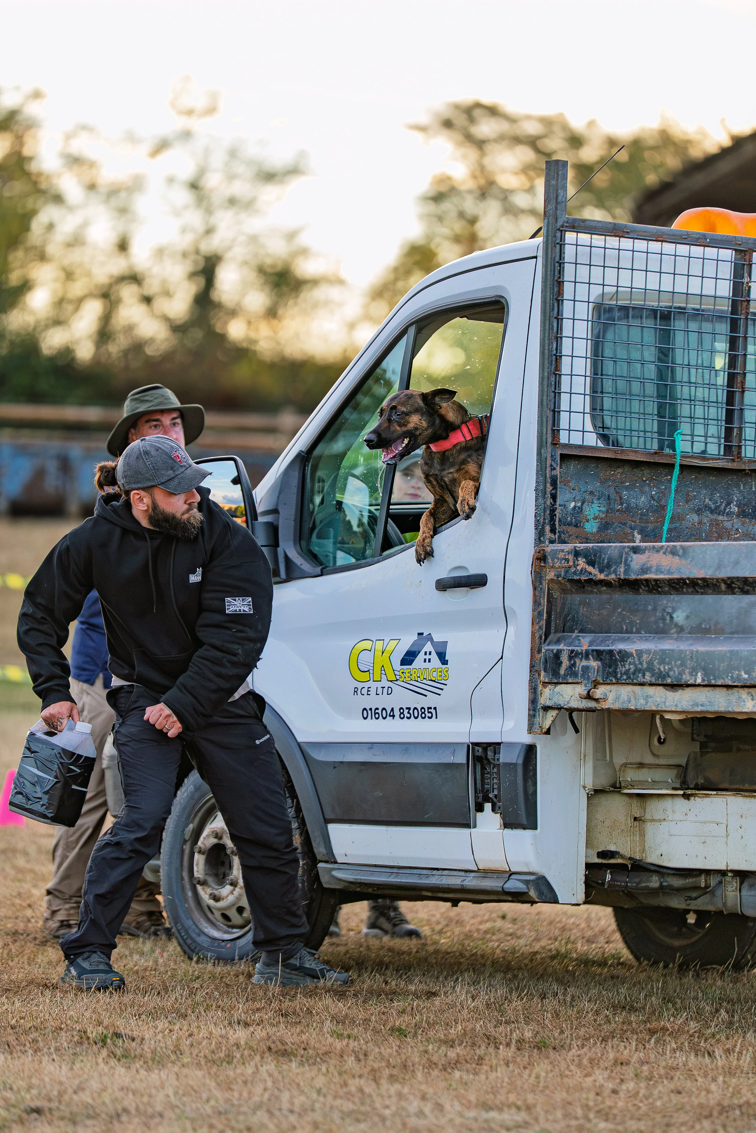 Two men, one wearing a gray hat and the other a baseball cap, are approaching a white utility truck with a muddy flatbed. A dog with a red collar is leaning out of the truck window, looking to the side. The background shows blurred trees and a fence 