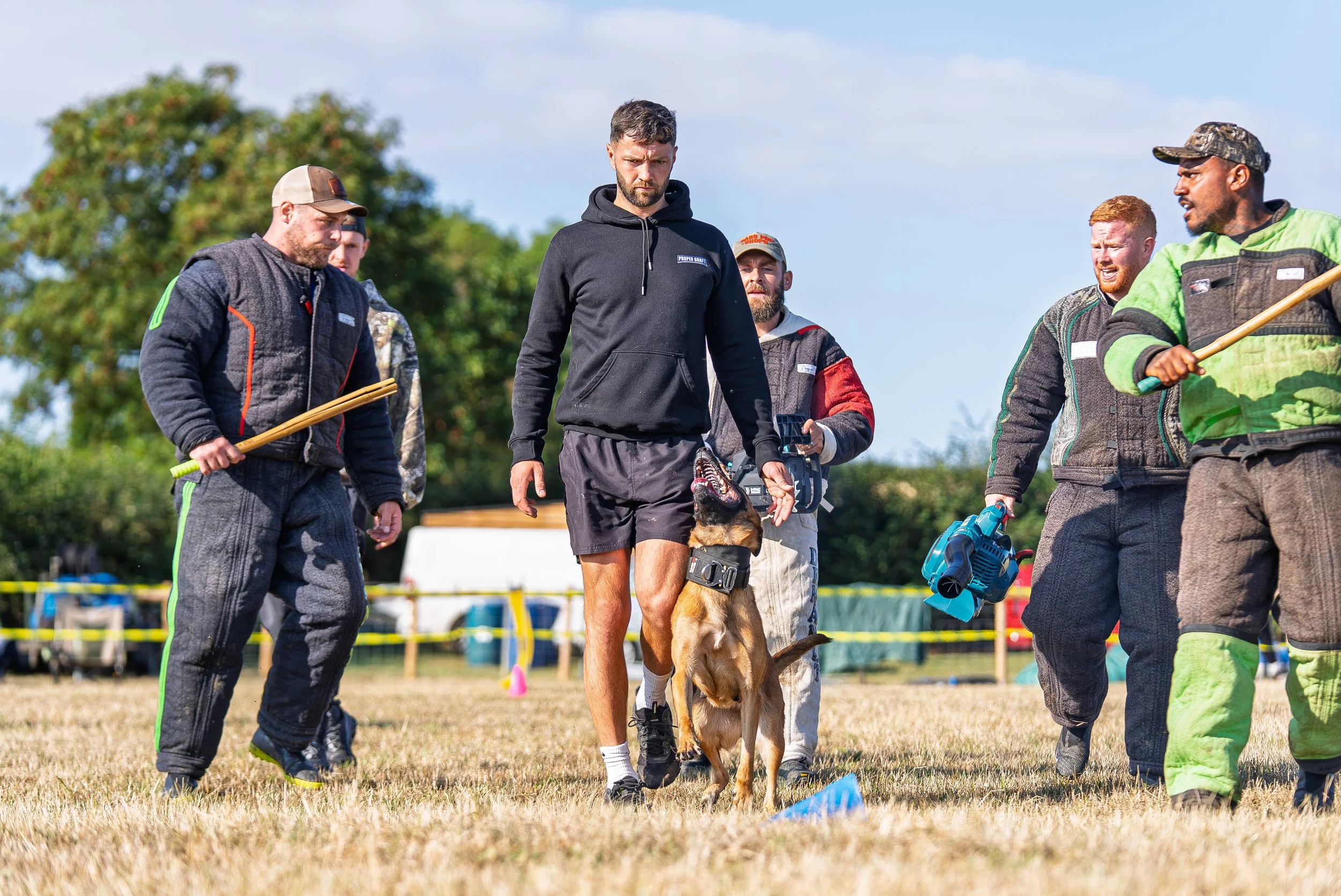 Group of people, including a man with a dog wearing a harness, participating in an outdoor dog training or agility event on a grassy field, with some spectators and equipment visible in the background.