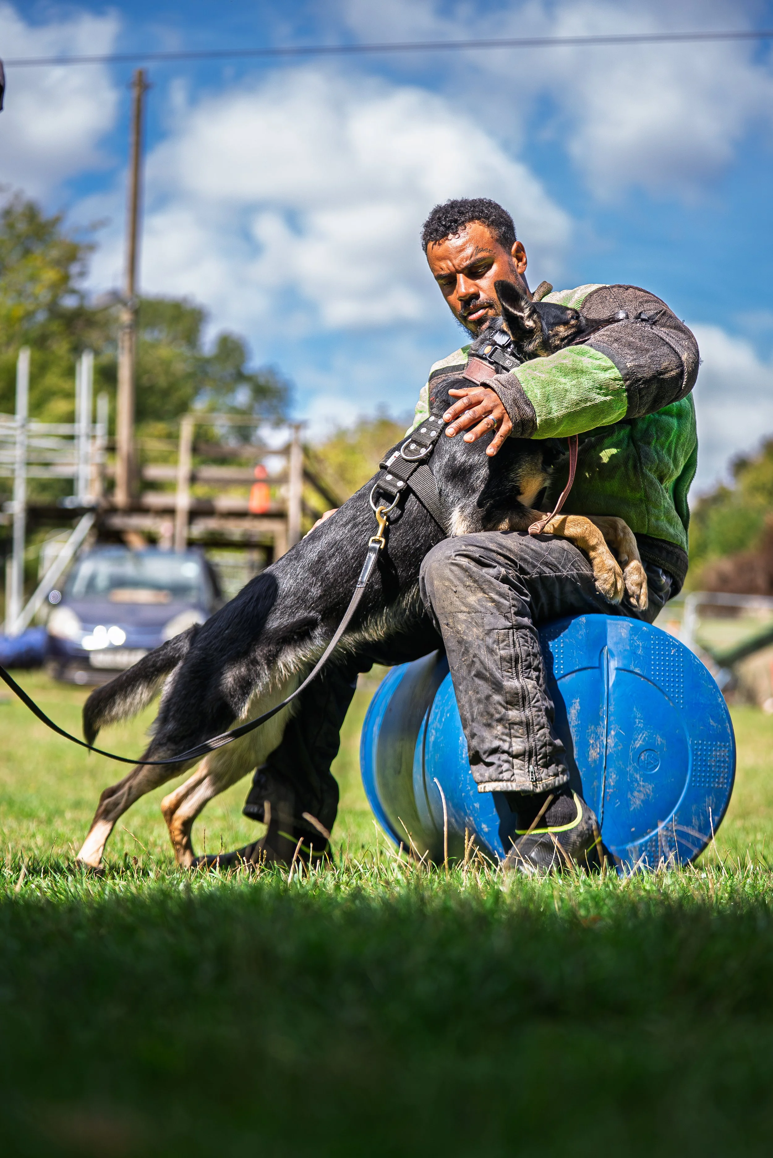 A man holding a dog during a training exercise outdoors, with a blue and gray tire obstacle on grass and a blue sky with clouds in the background.