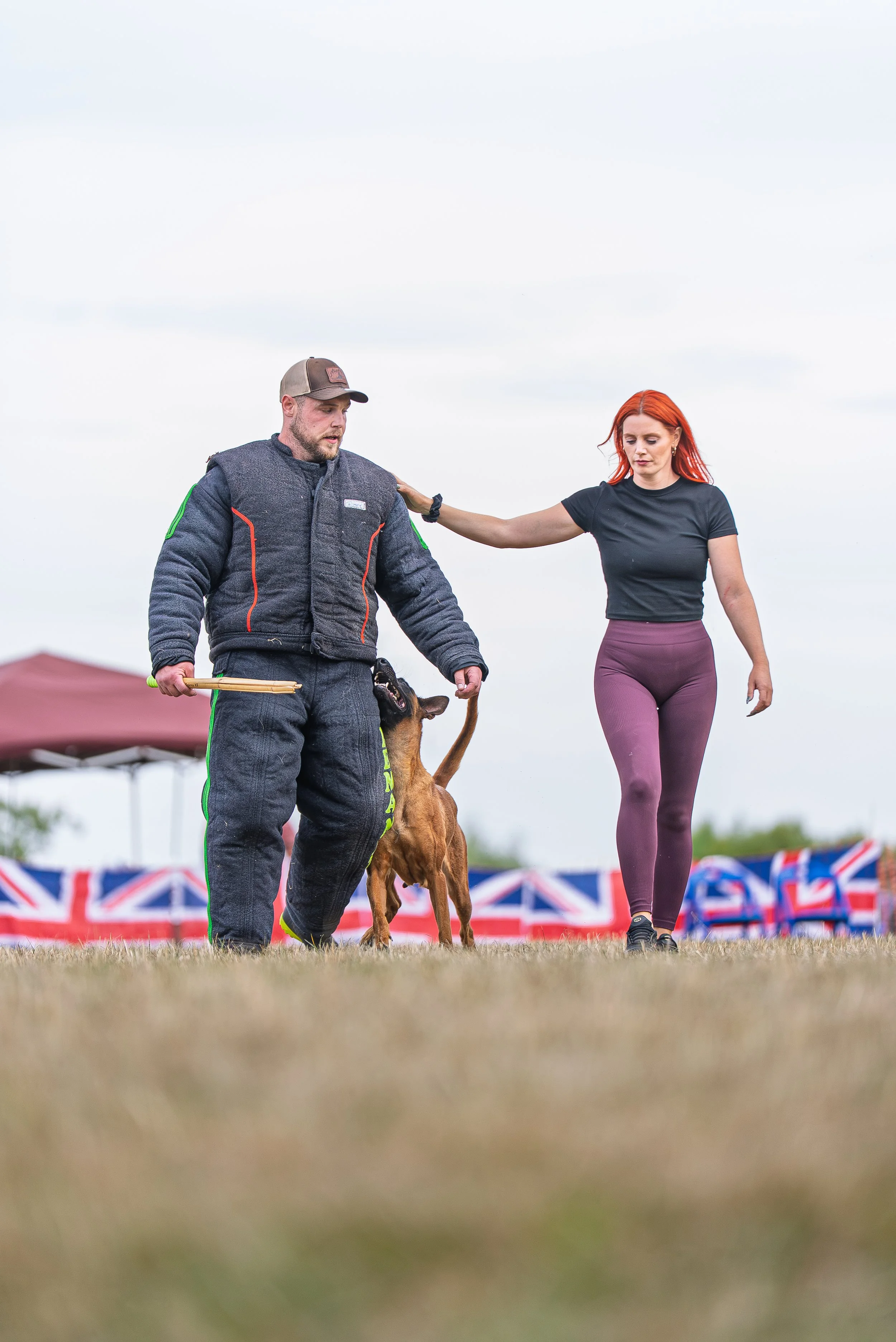 Man dressed in a black protective suit holds a dog by a leash as a woman stands nearby, touching the man's shoulder, during an outdoor event under cloudy skies.